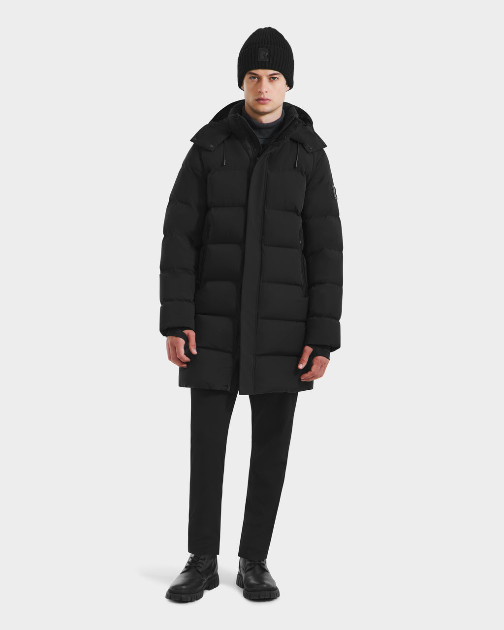 A person faces forward against a plain white background, wearing the Rudsak DAO - 6124604 Black mens mid-length puffer with a weather-resistant shell, paired with a black beanie, pants, and shoes.