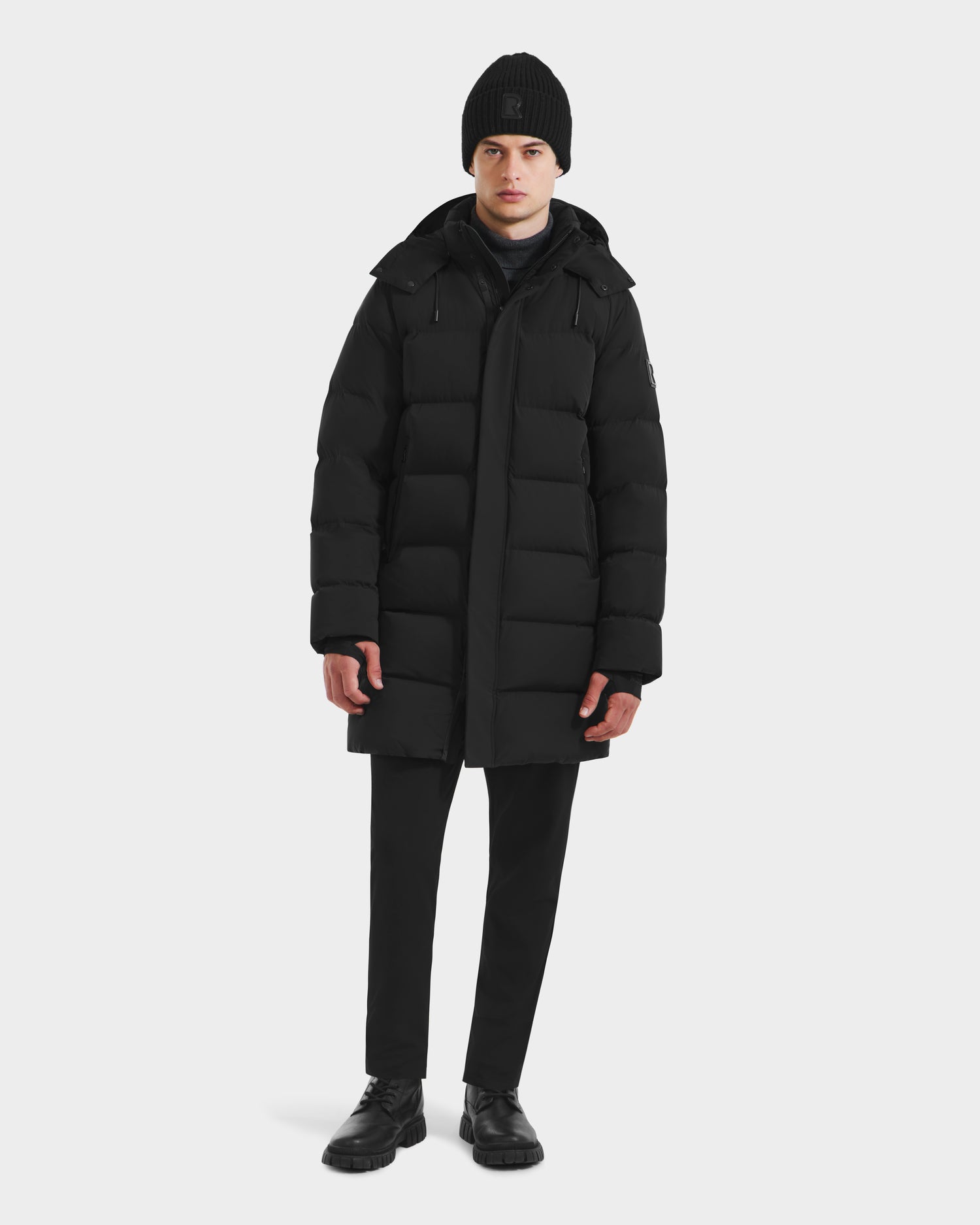 A person faces forward against a plain white background, wearing the Rudsak DAO - 6124604 Black mens mid-length puffer with a weather-resistant shell, paired with a black beanie, pants, and shoes.