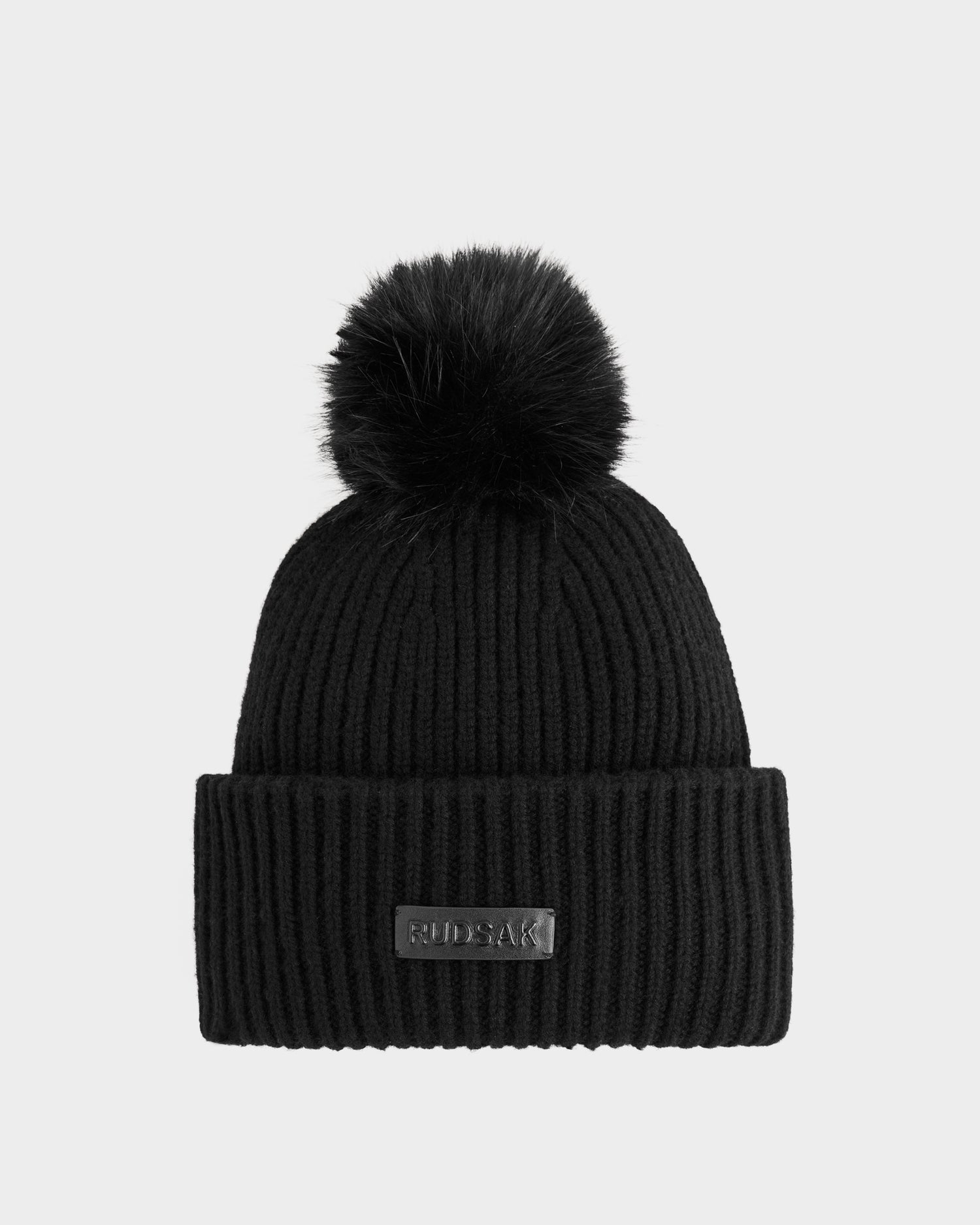The Rudsak DARA - 8425019 Black is a women’s ribbed knit tuque with a large faux fur pom pom, made from a soft cashmere wool blend and finished with a folded cuff featuring a rectangular patch label on the front.