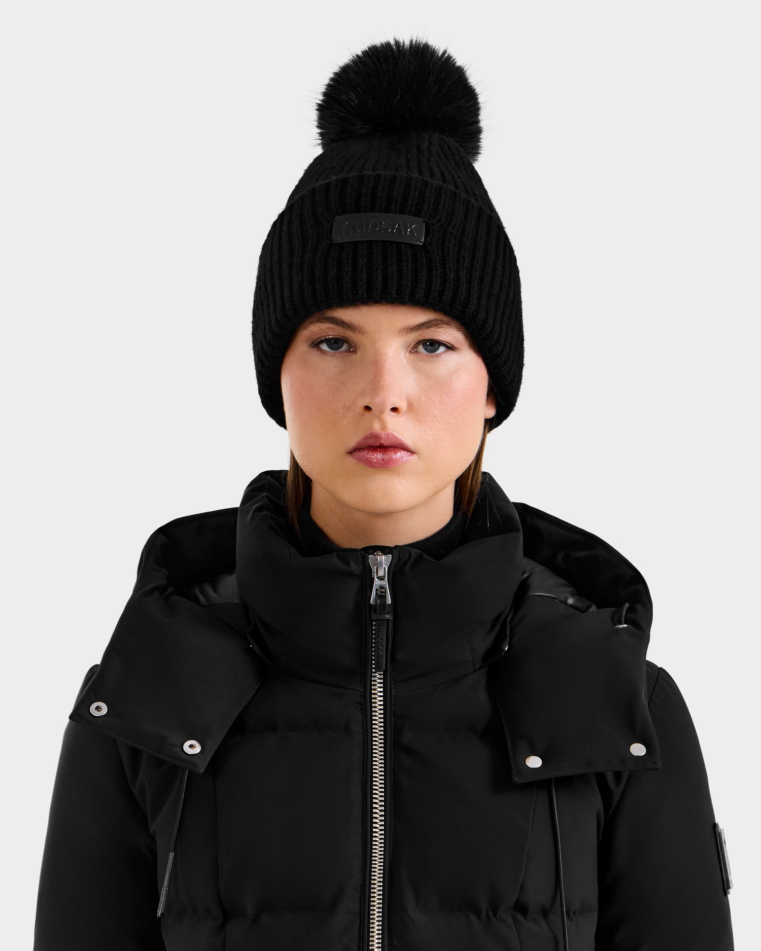 A woman faces the camera against a plain light background, wearing the Rudsak DARA - 8425019 Black cashmere wool tuque with faux fur pom pom and a black high-collar puffer jacket, her expression neutral.