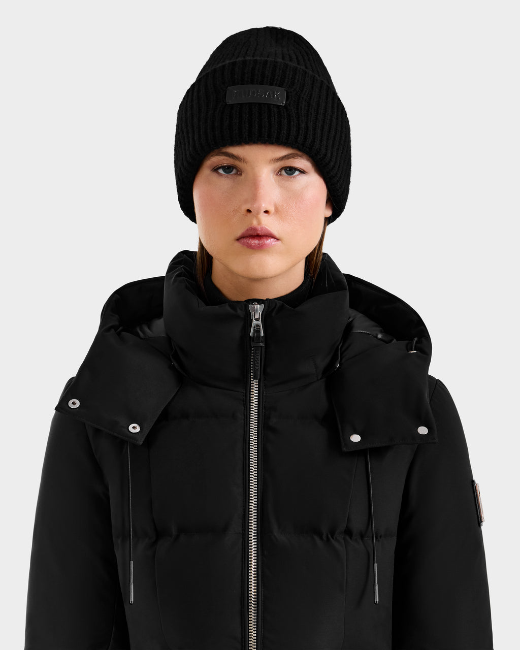 A woman wearing the Rudsak DARA - 8425019 Black cashmere tuque and a black puffer jacket faces forward with a neutral expression against a light gray background—a chic, cold-weather essential look.