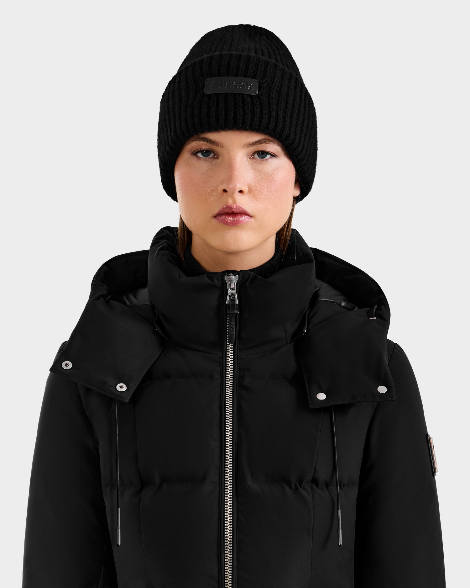 A woman wearing the Rudsak DARA - 8425019 Black cashmere tuque and a black puffer jacket faces forward with a neutral expression against a light gray background—a chic, cold-weather essential look.