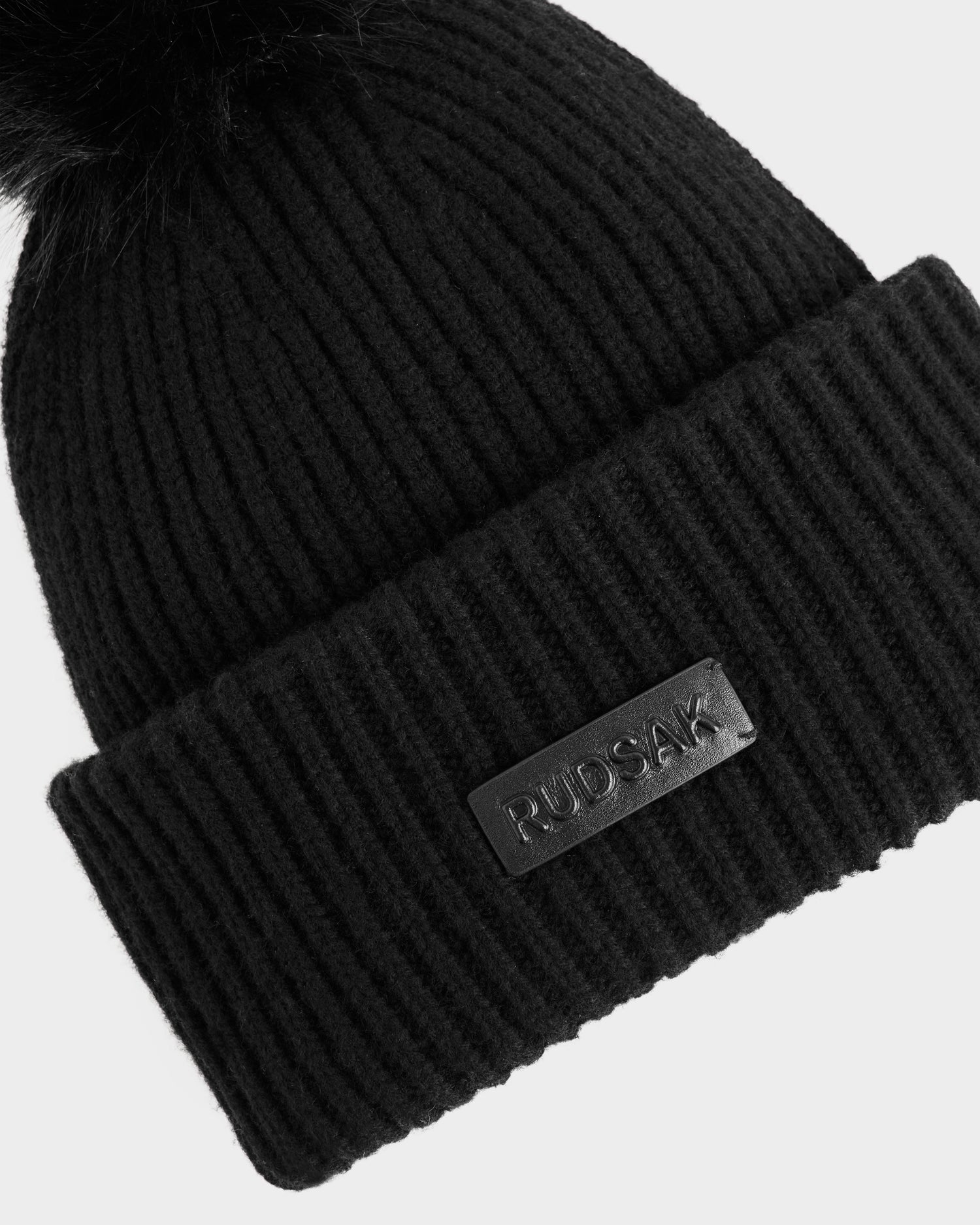 The DARA - 8425019 Black women's winter hat by Rudsak features a black ribbed knit, folded brim, Rudsak leather patch, and a black faux fur pom-pom, displayed on a white background.
