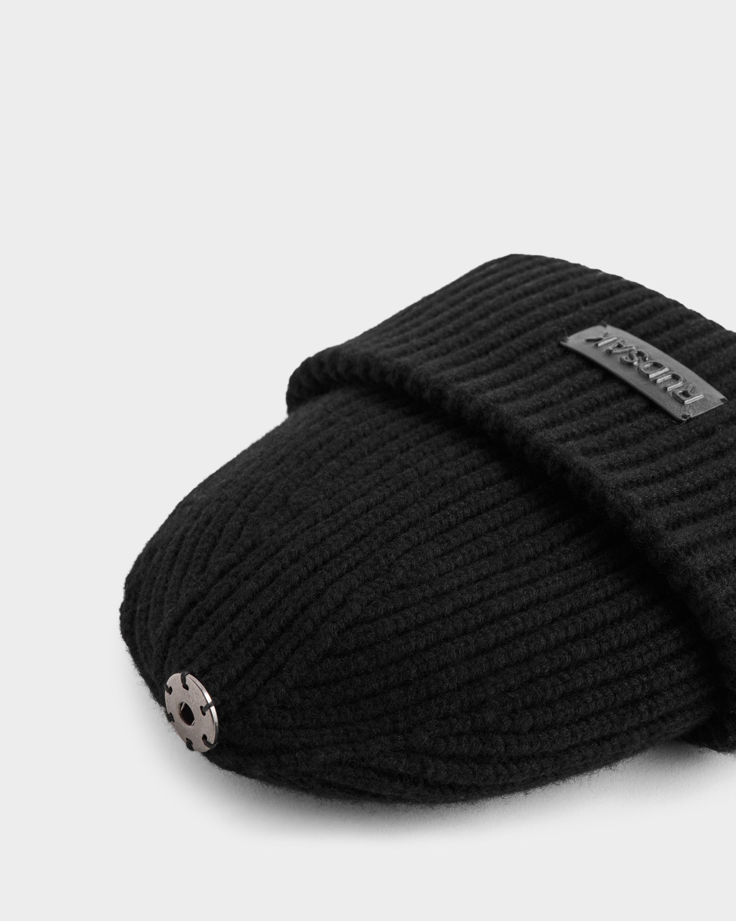 A close-up of the Rudsak DARA - 8425019 Black slipper, featuring black ribbed knit cashmere-wool blend, a metallic button on the toe, and a fabric label on the upper, shown against a white background.