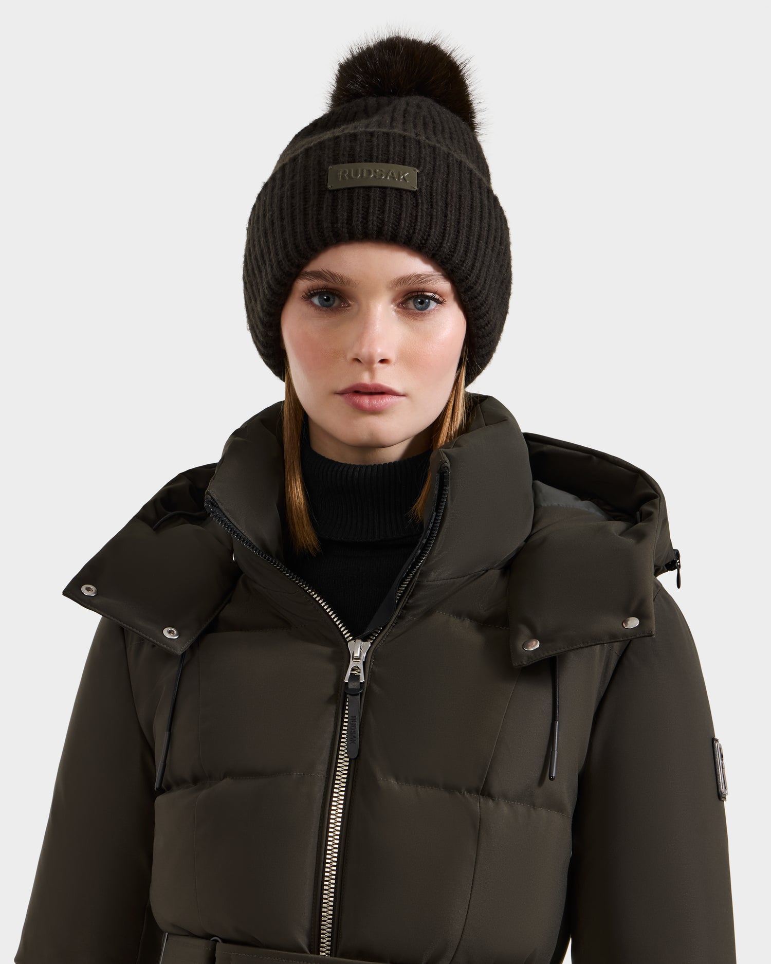 A woman wears the Rudsak DARA - 8425019 dark olive puffer jacket and a matching women’s tuque with faux fur pom pom, standing against a plain light gray background.