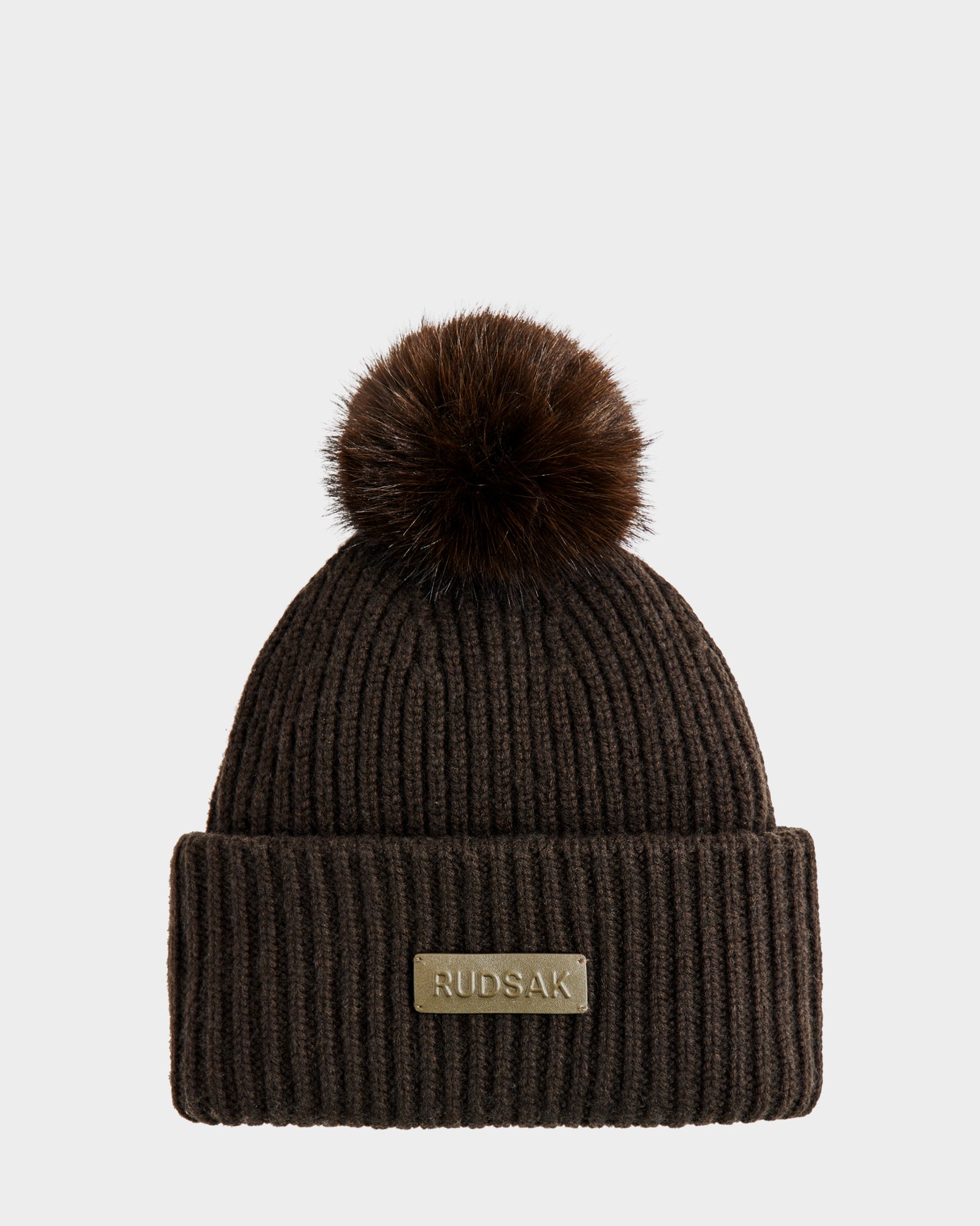 The Rudsak DARA - 8425019 is a dark olive ribbed women’s tuque with a large faux fur pom pom, a cashmere wool blend, and a folded brim featuring a rectangular RUDSAK label. Shown on a plain light gray background.