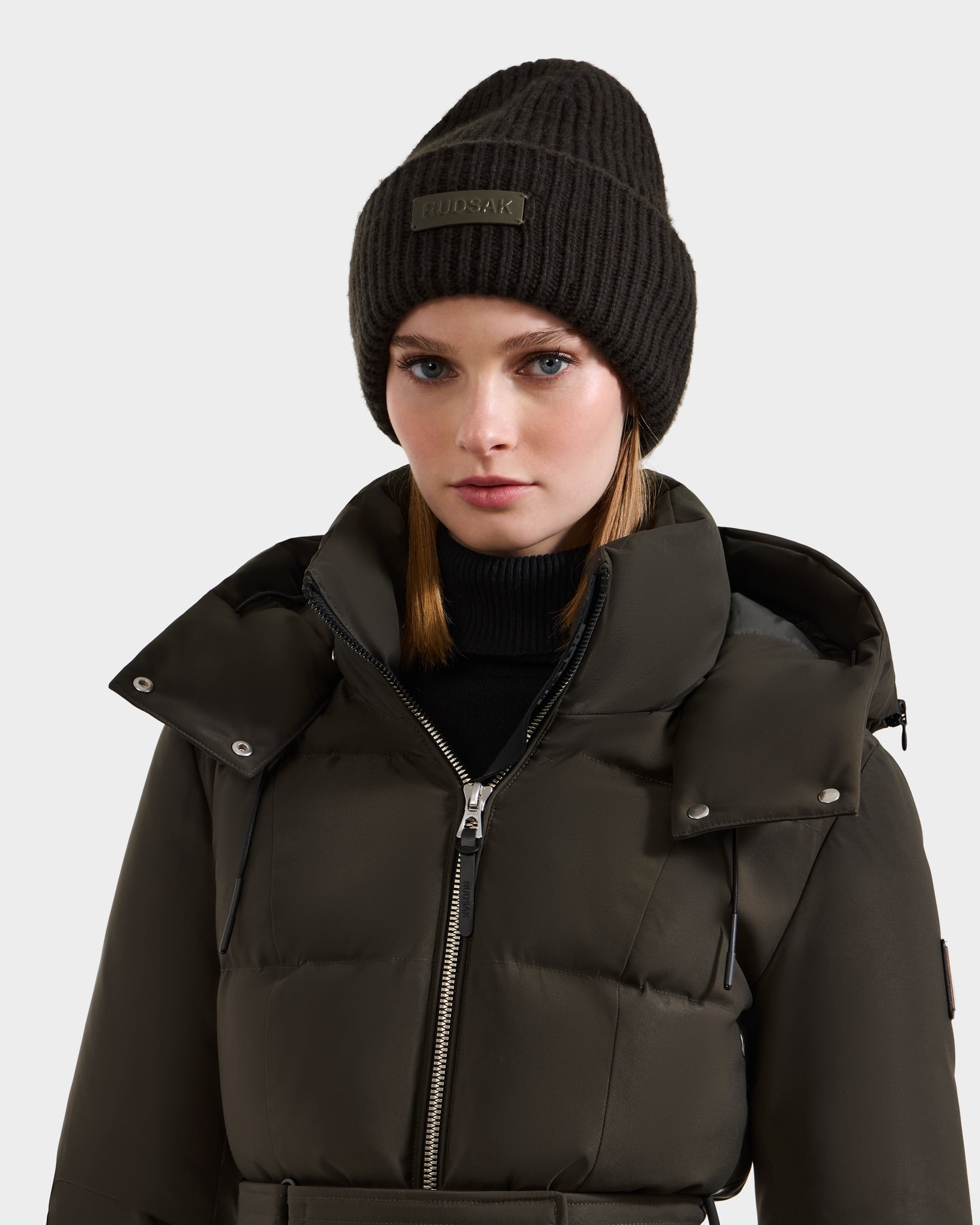 A woman in the Rudsak DARA - 8425019 puffer jacket in dark olive pairs it with a women’s tuque featuring a faux fur pom pom, standing against a plain light grey background.