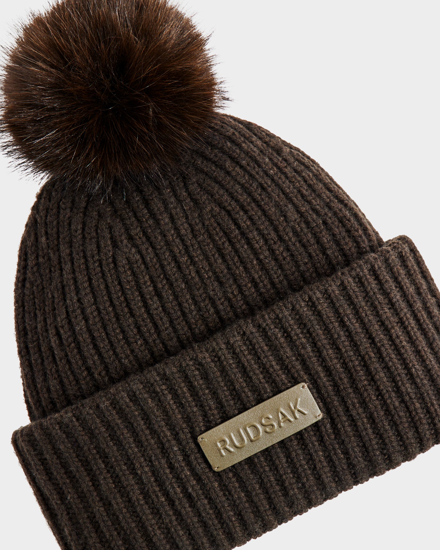 The DARA - 8425019 by Rudsak is a ribbed, dark olive women’s knit beanie with a large faux fur pom pom, crafted from a cashmere wool blend and finished with a folded brim and metal Rudsak logo plate.