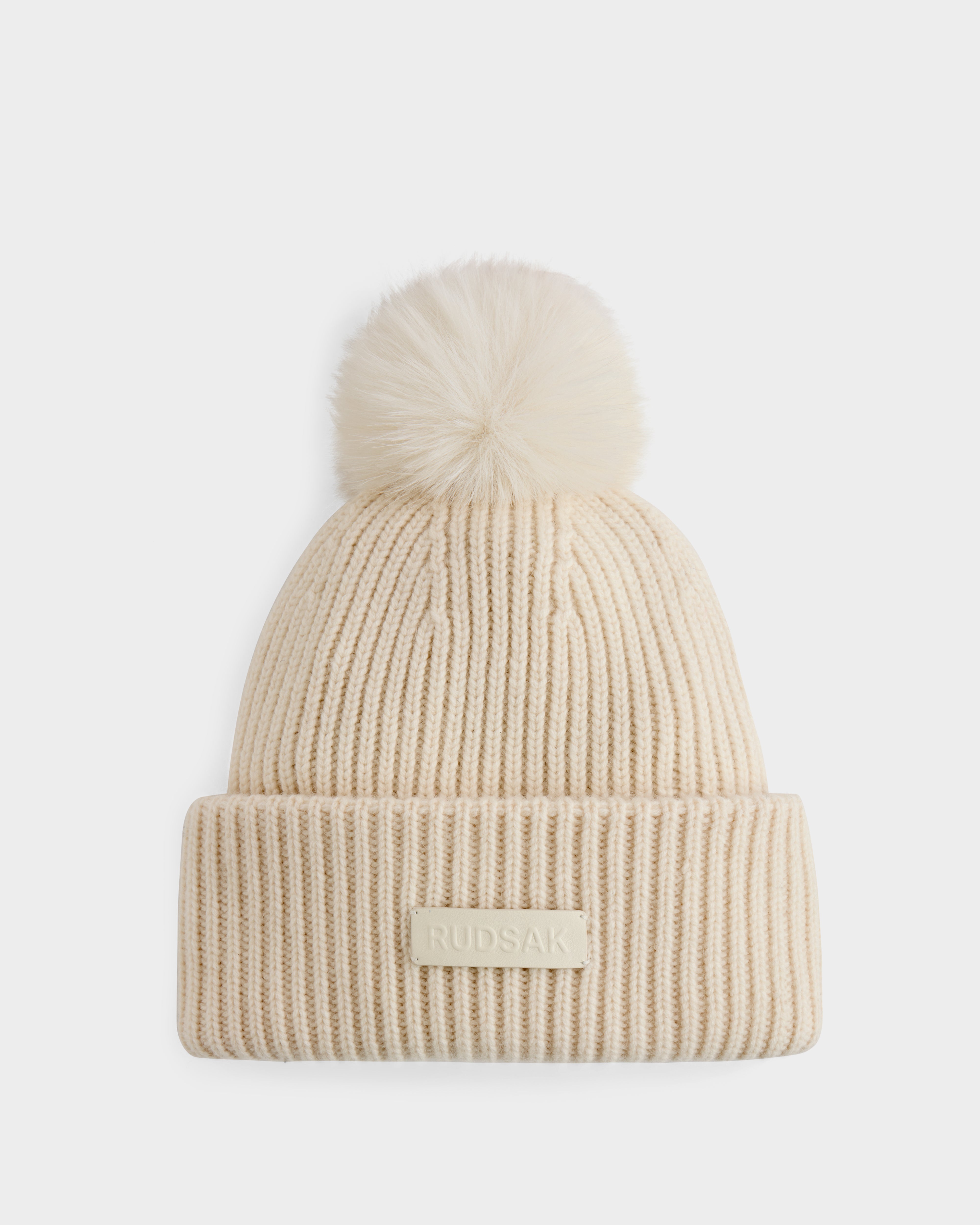 The DARA - 8425019 Vanilla by Rudsak is a cream ribbed knit women’s tuque featuring a large faux fur pom pom, crafted from a cashmere wool blend, with a folded brim and rectangular RUDSAK patch.