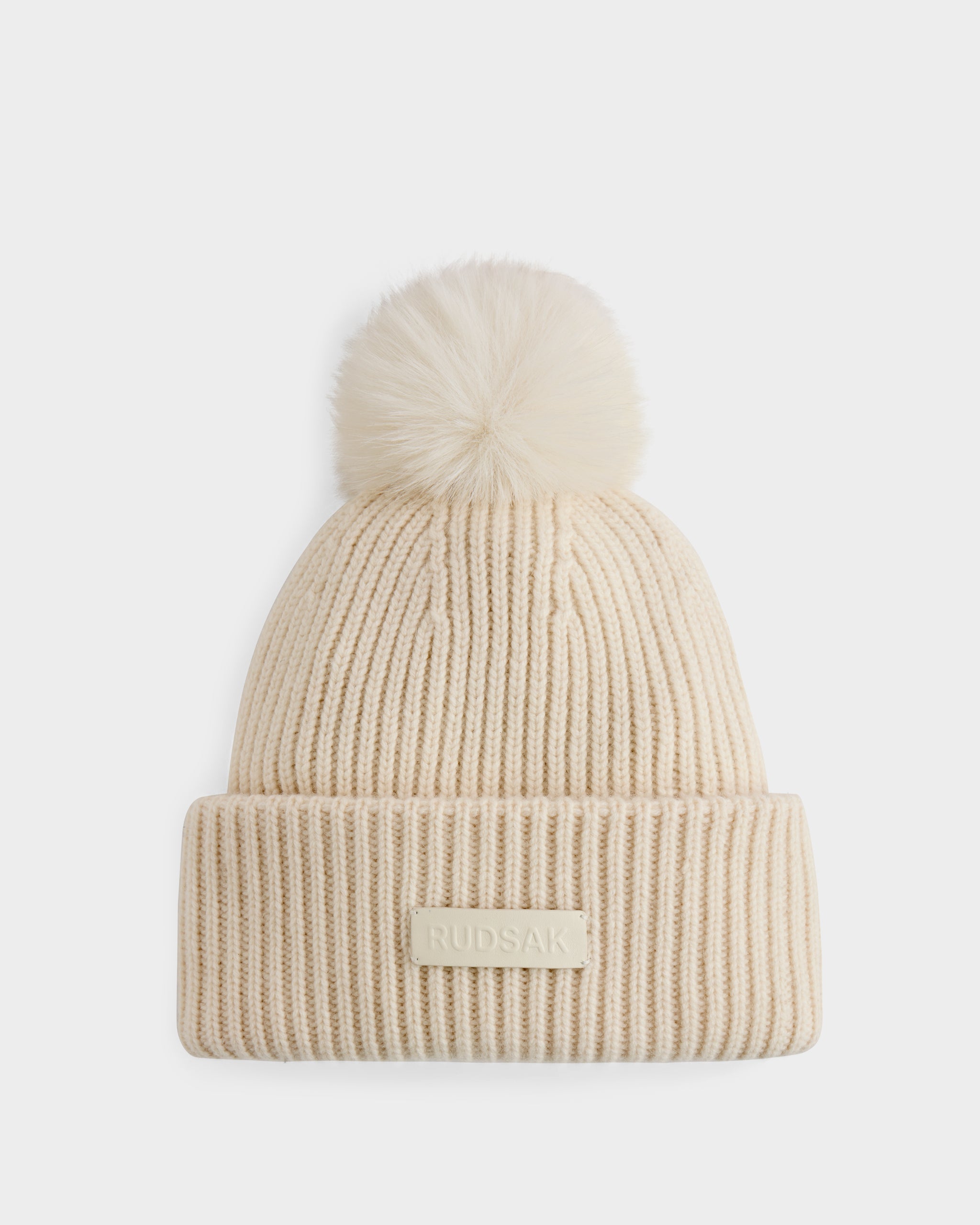 The DARA - 8425019 Vanilla by Rudsak is a cream ribbed knit women’s tuque featuring a large faux fur pom pom, crafted from a cashmere wool blend, with a folded brim and rectangular RUDSAK patch.