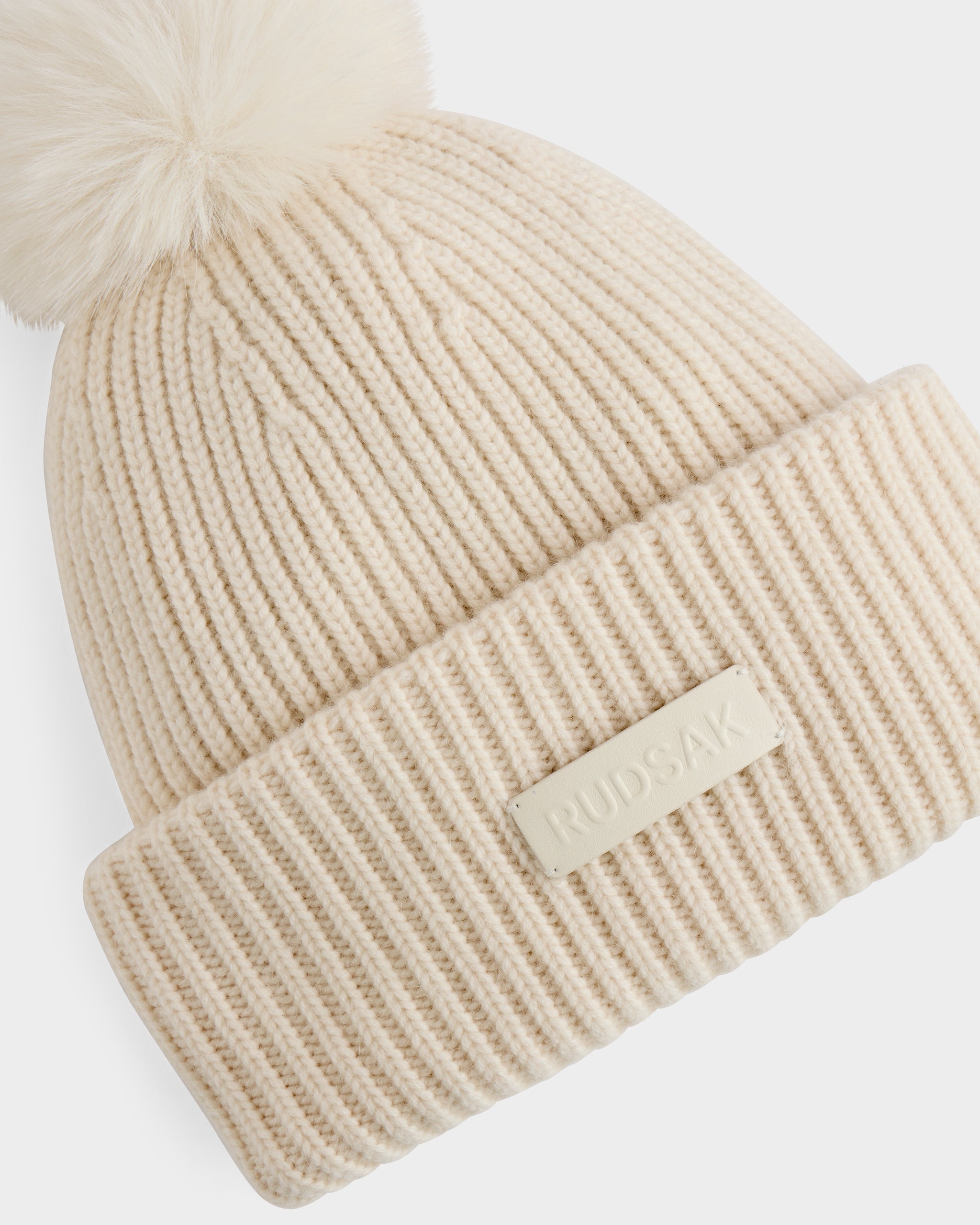 The DARA - 8425019 Vanilla by Rudsak is an ivory ribbed knit women’s tuque with a large faux fur pom pom, crafted from a soft cashmere wool blend and finished with a folded brim featuring the Rudsak patch.