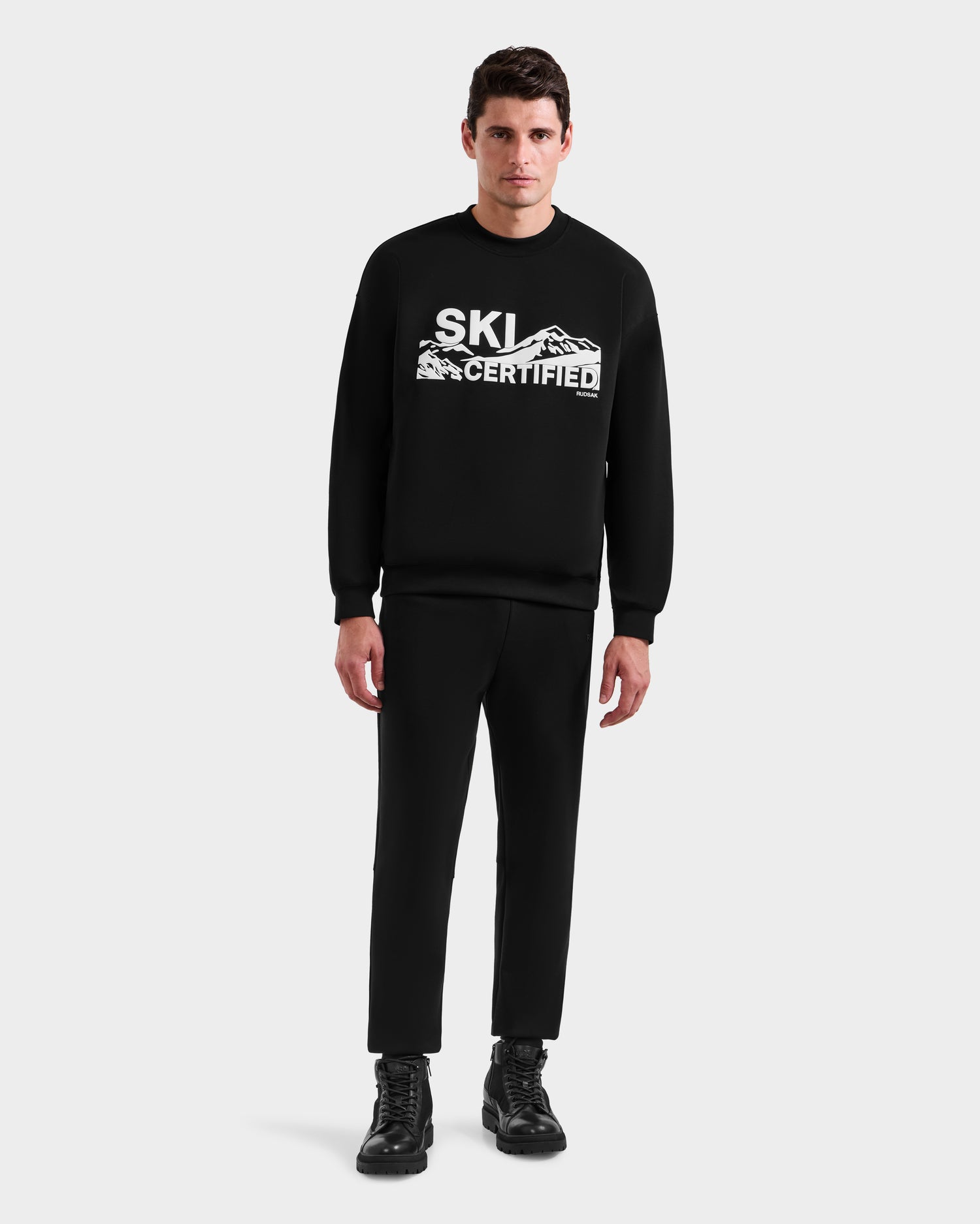 A man stands facing forward, wearing Rudsak's DECLAN - 6525648 Black lace-up boots with a ski certified sweatshirt, black pants, and a mountain graphic, set against a plain light background.