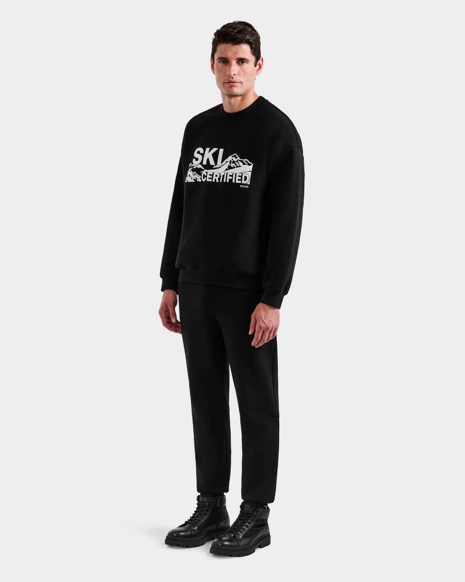 A man stands facing forward in a Rudsak DECLAN - 6525648 Black sweatshirt with mountain graphics, paired with black pants and boots against a plain light gray background.