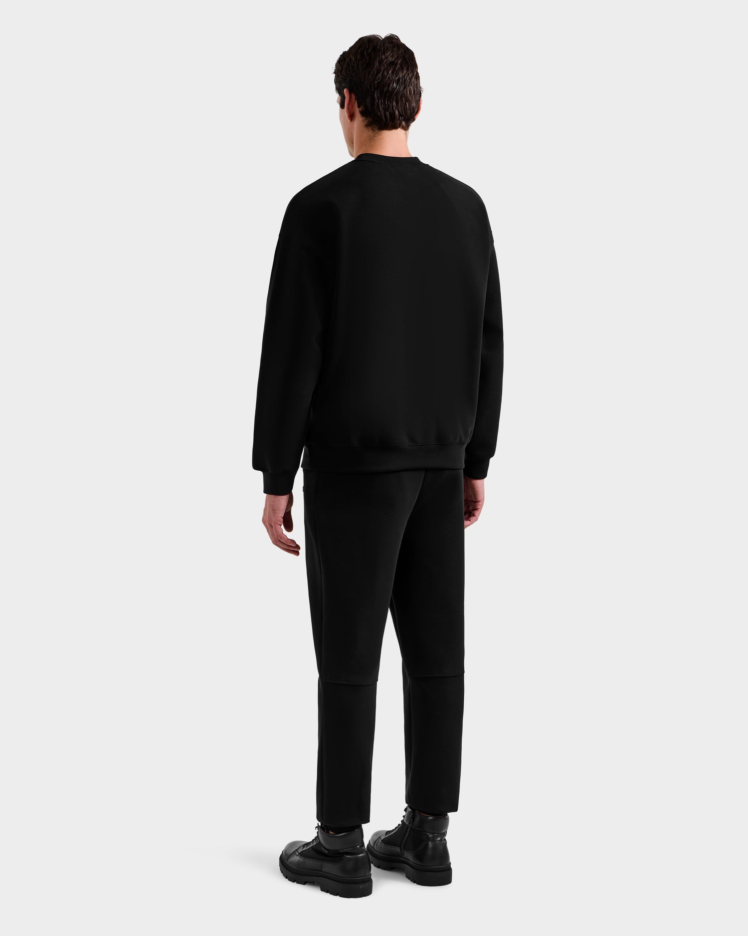 A man faces away from the camera, wearing the Rudsak DECLAN - 6525648 Black crewneck sweatshirt, paired with black pants and boots, set against a plain light gray background.