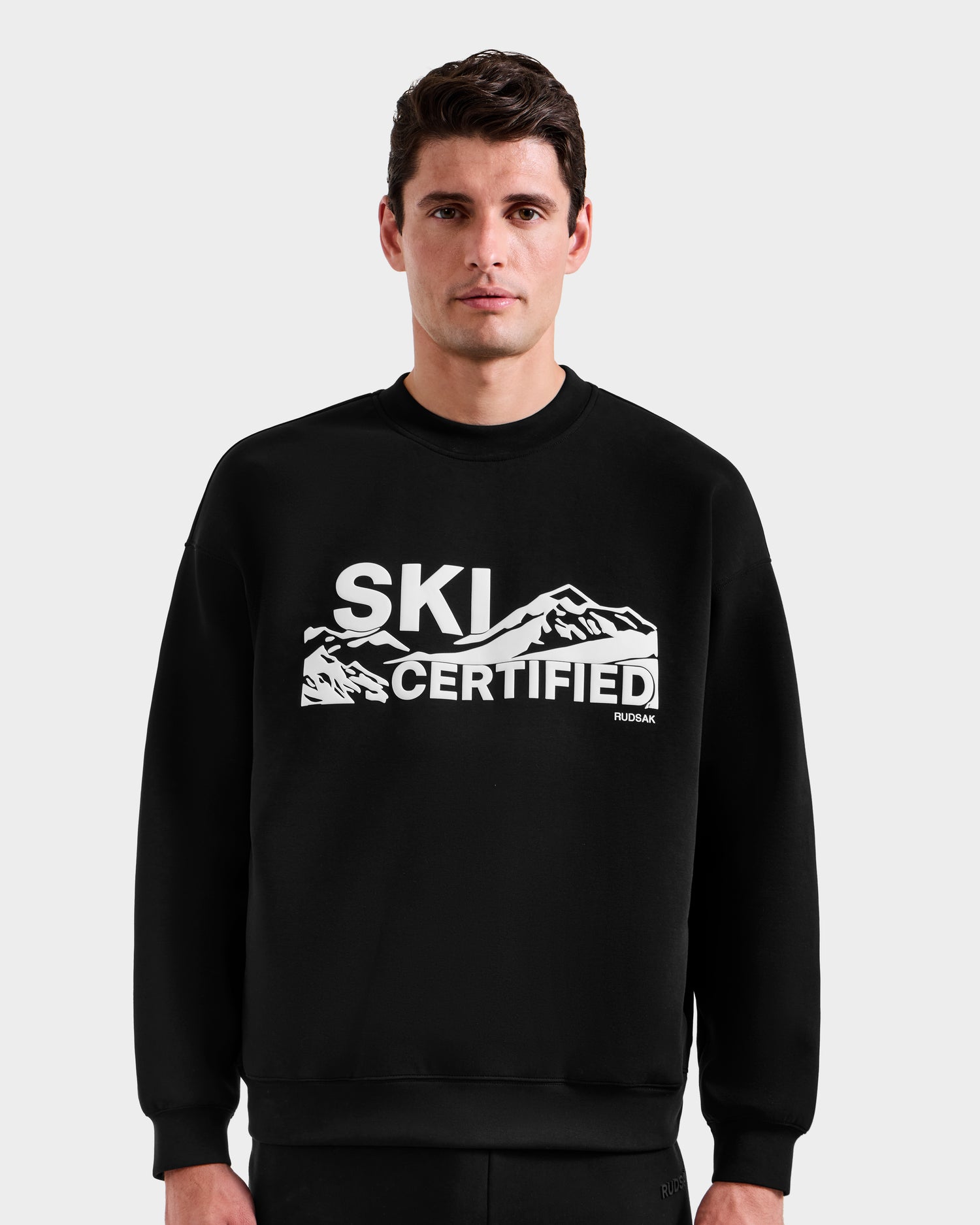 A man wears the Rudsak DECLAN - 6525648 Black men’s crewneck sweatshirt with a white mountain graphic and bold “SKI CERTIFIED” text, standing against a plain, light-colored background.