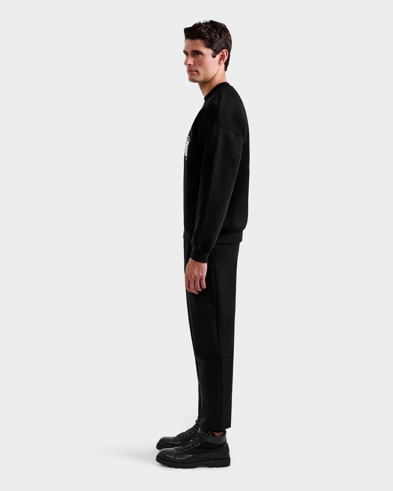 A man stands in profile against a plain white background, wearing the Rudsak DECLAN - 6525648 Black men’s crewneck sweatshirt with matching black pants and shoes.