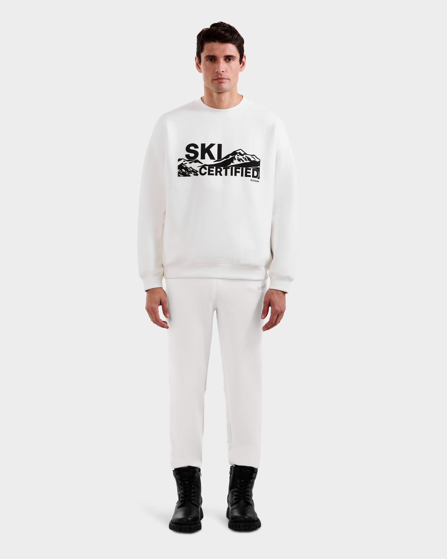 A man faces forward wearing the Rudsak DECLAN - 6525648 Iceland men’s crewneck sweatshirt with SKI CERTIFIED and mountain graphics, paired with white pants and black boots, against a plain light background.