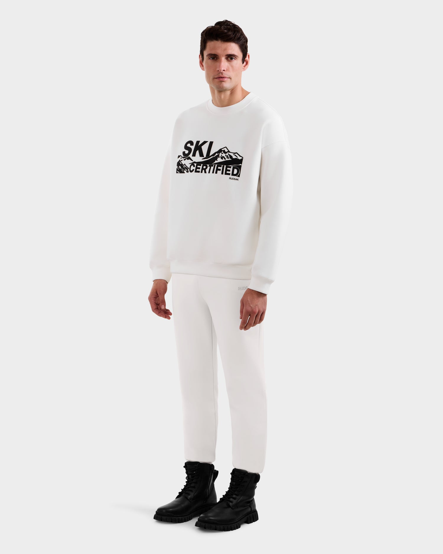 A man faces forward wearing the Rudsak DECLAN - 6525648 Iceland men’s crewneck sweatshirt with “SKI CERTIFIED” and a mountain graphic, paired with white pants and black boots, set against a plain white background.