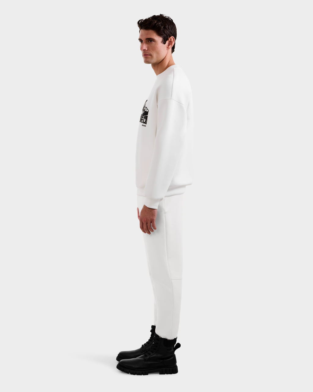 A man stands in profile against a plain white background wearing Rudsak's DECLAN - 6525648 Iceland crewneck sweatshirt and matching all-white sweatpants set, styled with black lace-up boots.