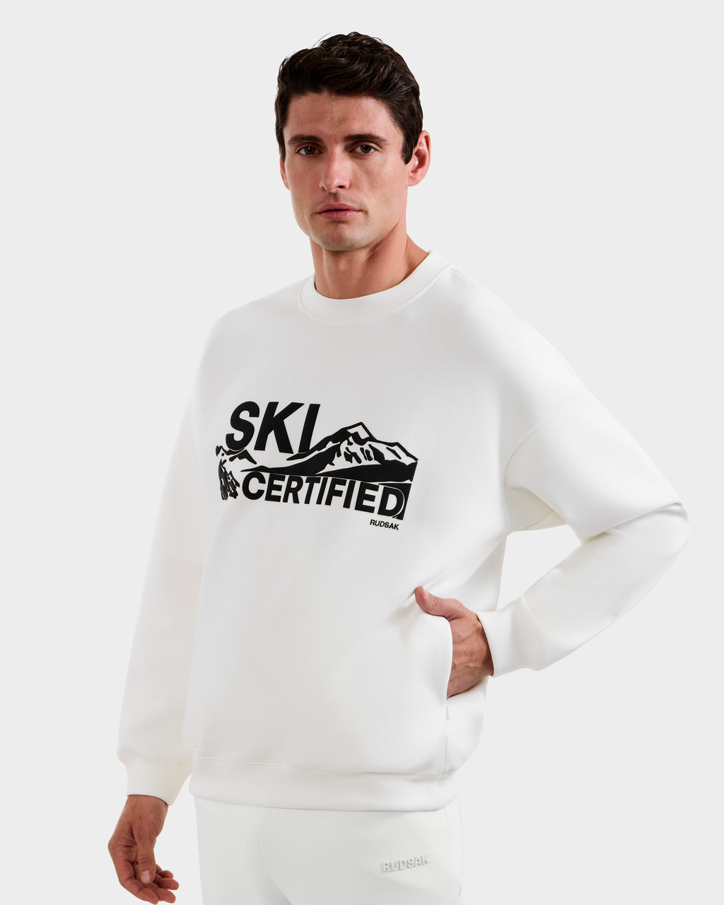A man wears the Rudsak DECLAN - 6525648 Iceland men’s crewneck sweatshirt featuring "SKI CERTIFIED" and a bold black mountain graphic on the front, standing against a plain light background.