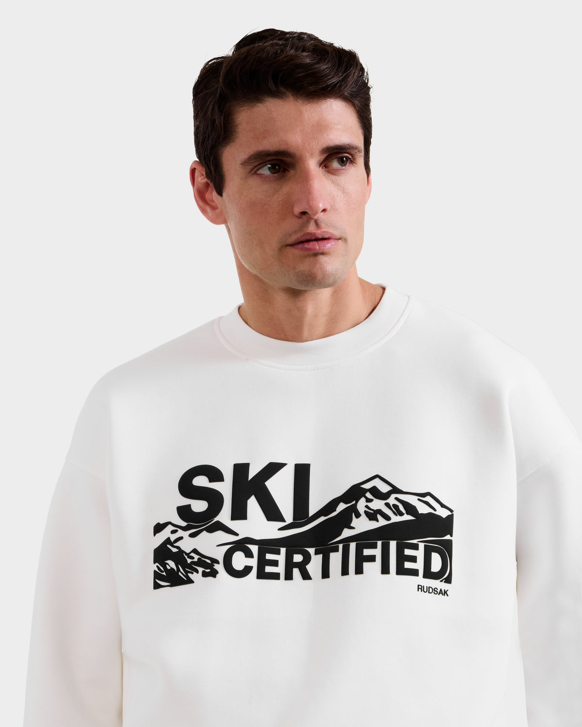Model wears the Rudsak DECLAN - 6525648 Iceland men’s crewneck sweatshirt, featuring a black mountain graphic and "SKI CERTIFIED" text on the front, turning slightly to his right to highlight this stylish piece.