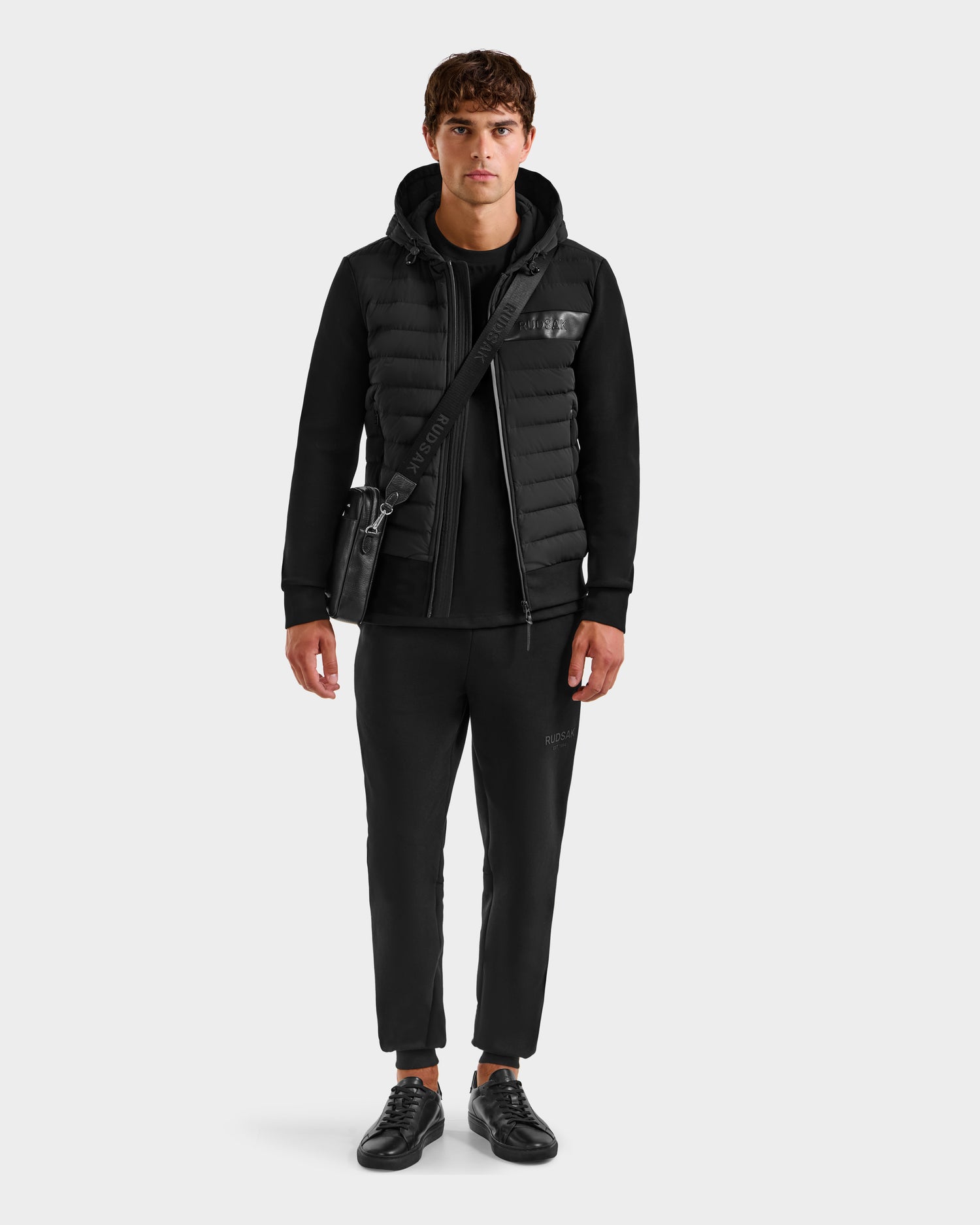 A young man faces forward against a light gray background, wearing the Rudsak DEE MP - 6125673 Black men’s lightweight down jacket with RDS-certified duck down, paired with a black T-shirt, pants, sneakers, and a crossbody bag.