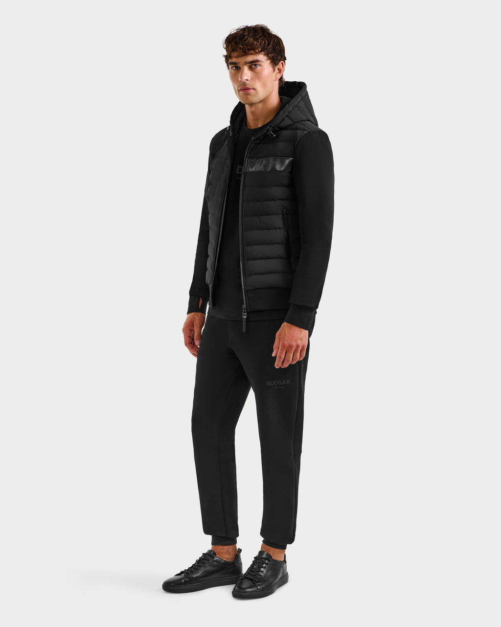 A young man faces slightly left wearing the Rudsak DEE MP - 6125673 Black men’s lightweight down jacket with RDS-certified duck down, paired with a black sweatshirt, jogger pants, and sneakers against a plain light gray background.