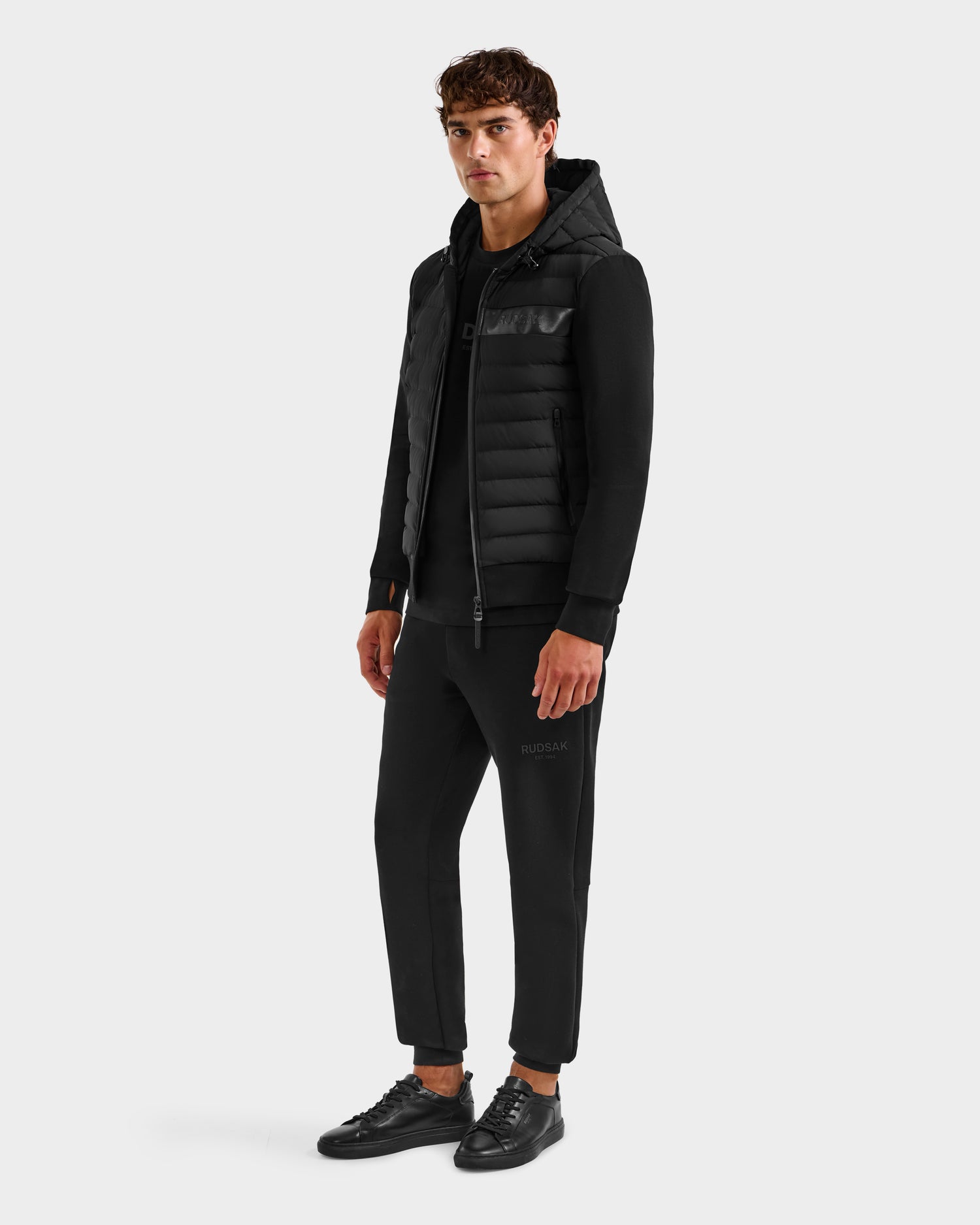 A young man faces slightly left wearing the Rudsak DEE MP - 6125673 Black men’s lightweight down jacket with RDS-certified duck down, paired with a black sweatshirt, jogger pants, and sneakers against a plain light gray background.