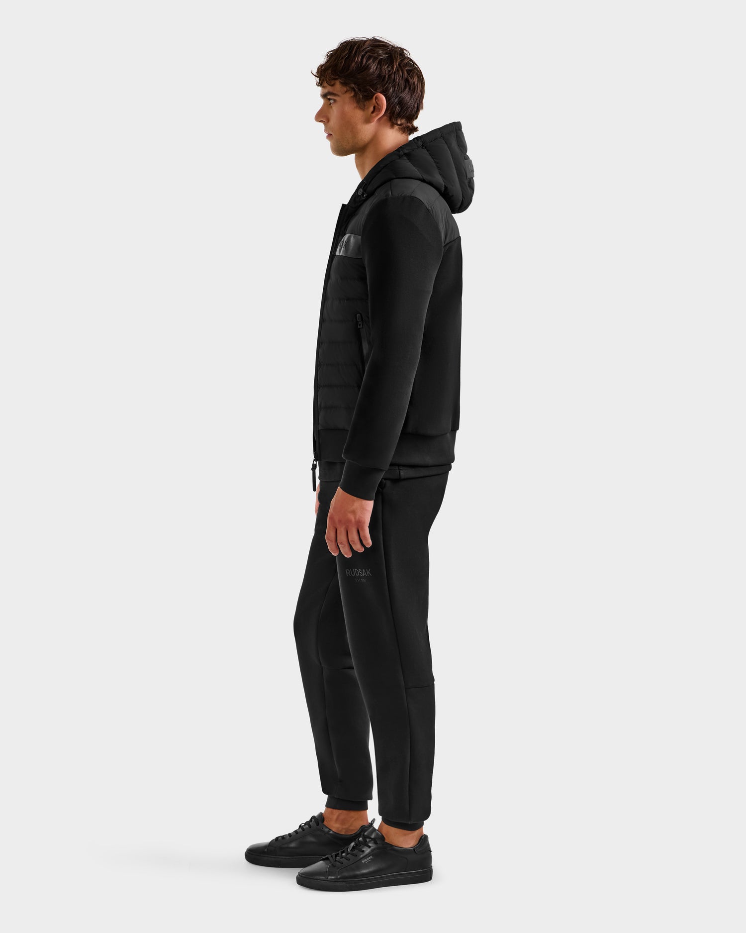 A man in profile wears the Rudsak DEE MP - 6125673 Black men’s lightweight down jacket with matching black joggers and sneakers against a light gray background—a perfect transitional staple.