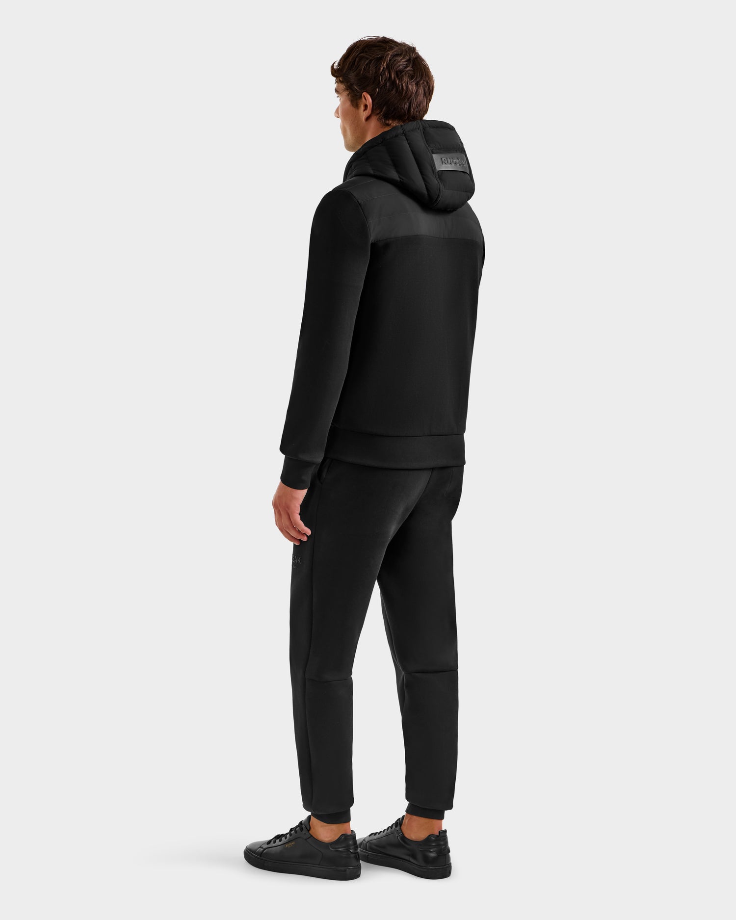 A person faces away, wearing the Rudsak DEE MP - 6125673 Black men’s lightweight down jacket—an ideal transitional staple—paired with black pants and sneakers, against a plain light gray background.
