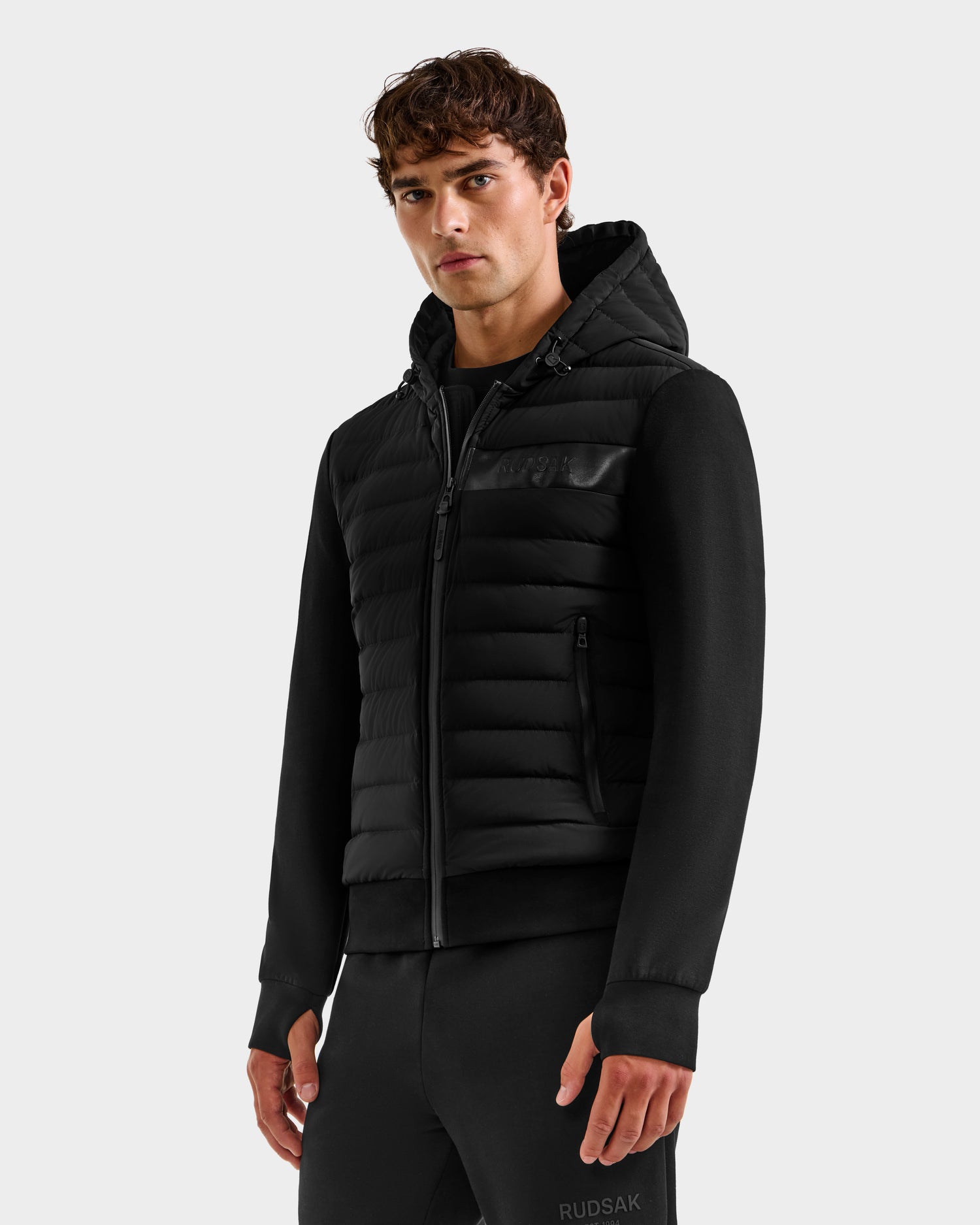 A young man models the Rudsak DEE MP - 6125673 Black men’s lightweight down jacket with zippered pockets and matching black pants against a light gray backdrop, highlighting this versatile transitional piece.