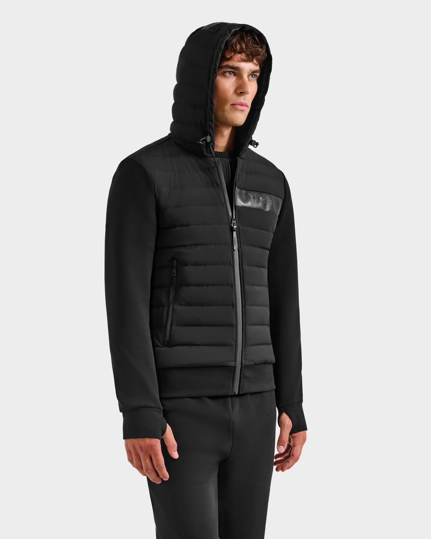 A man in the Rudsak DEE MP - 6125673 Black men’s lightweight down jacket with quilted front and sleeves stands against a plain light gray background, exuding urban polish.