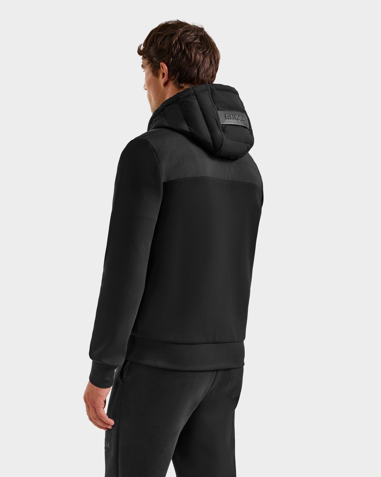 A man stands facing away, showcasing the Rudsak DEE MP - 6125673 Black hoodie with a puffer-style, RDS-certified duck down hood and matching sweatpants against a plain light backdrop.