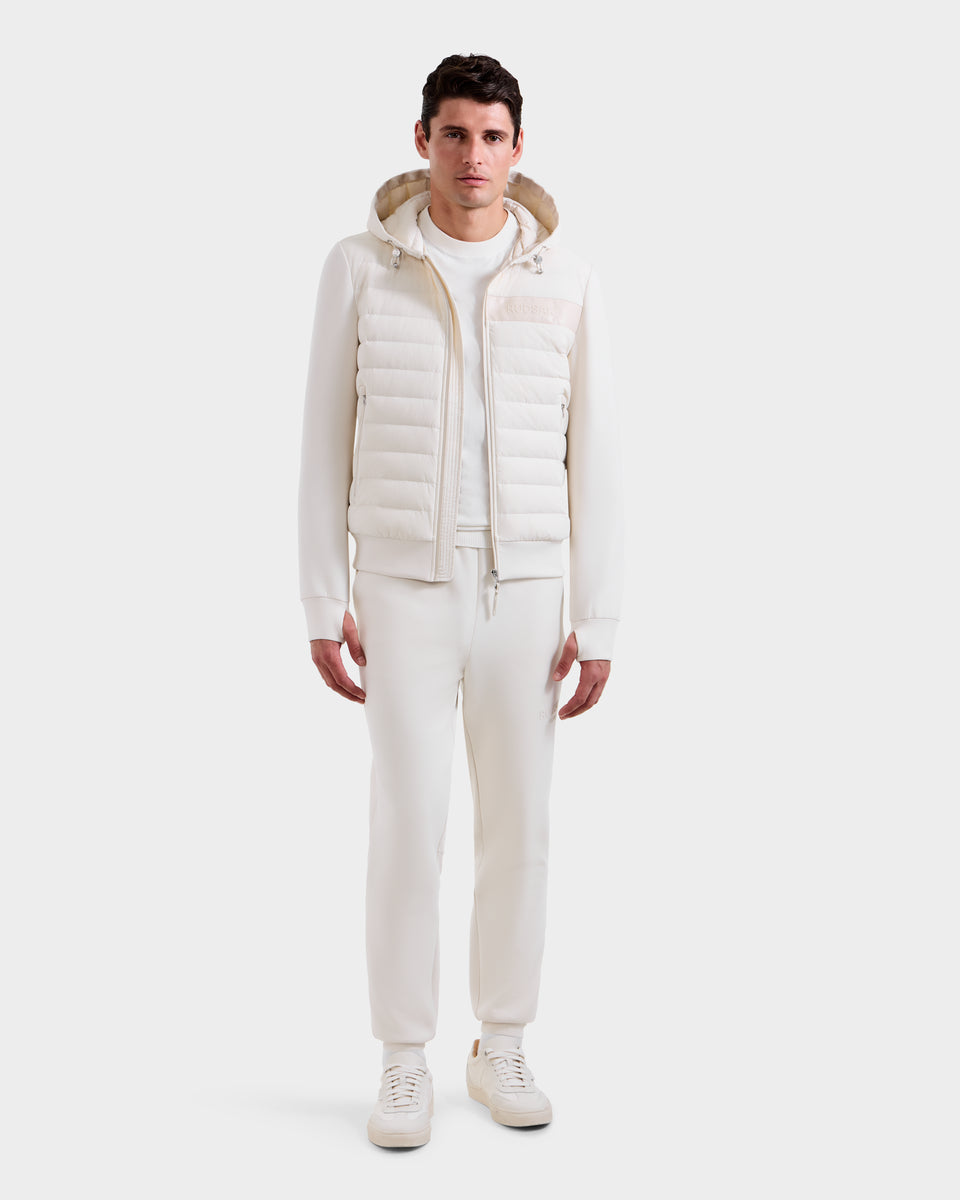 A man stands facing forward against a plain white background, wearing the Rudsak DEE MP - 6125673 Vanilla men’s lightweight down jacket filled with RDS-certified duck down, paired with a white sweater, pants, and sneakers.