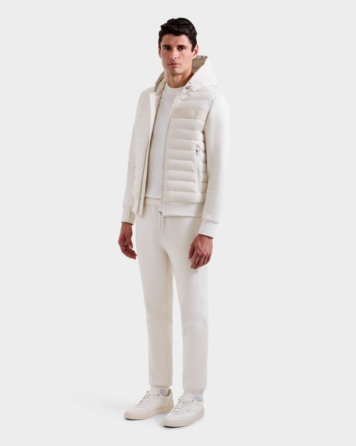 A man faces forward against a plain white background, wearing the Rudsak DEE MP - 6125673 Vanilla men’s lightweight down jacket with waterproof zippers, paired with matching pants, sweatshirt, and sneakers.