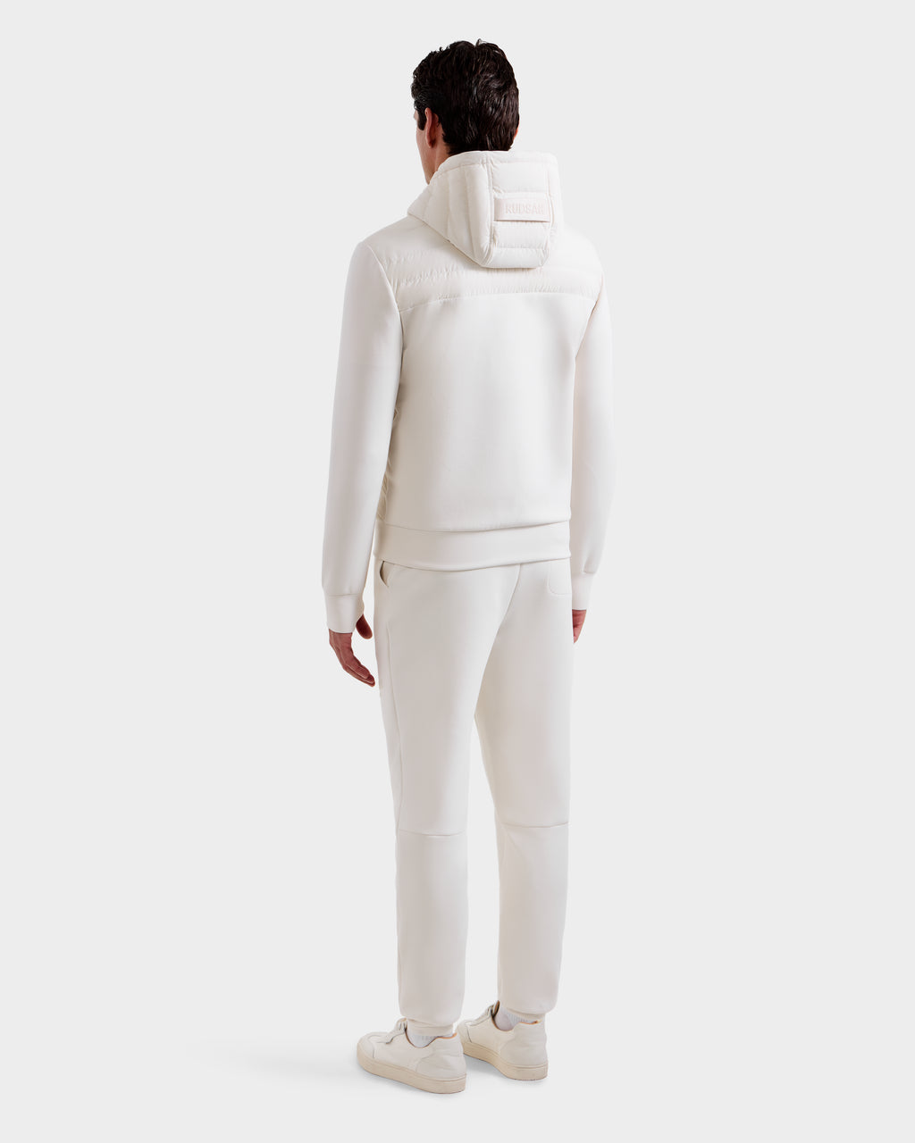 A man faces away from the camera, wearing Rudsak’s DEE MP - 6125673 Vanilla lightweight down jacket with waterproof zippers, matching pants, and white sneakers, all against a plain white background.