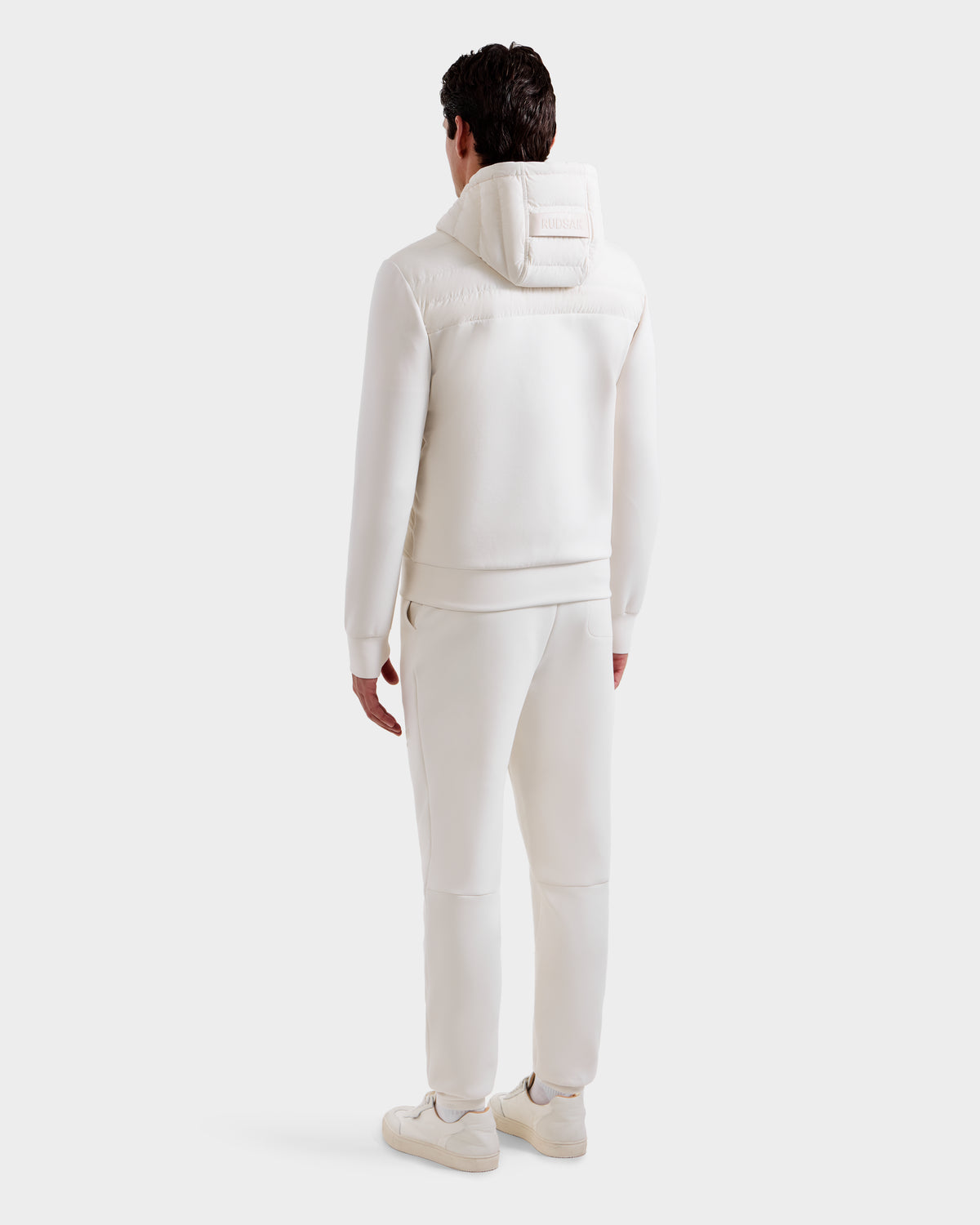 A man faces away from the camera, wearing Rudsak’s DEE MP - 6125673 Vanilla lightweight down jacket with waterproof zippers, matching pants, and white sneakers, all against a plain white background.