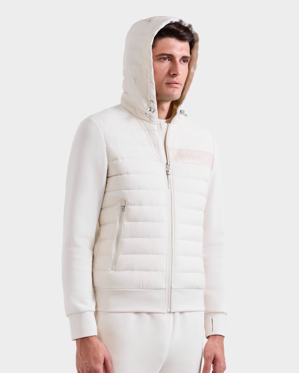 A man wearing a white Rudsak DEE MP - 6125673 Vanilla men’s lightweight down jacket with quilted padding and waterproof zippers stands against a plain background, looking slightly off to the side with a neutral expression.