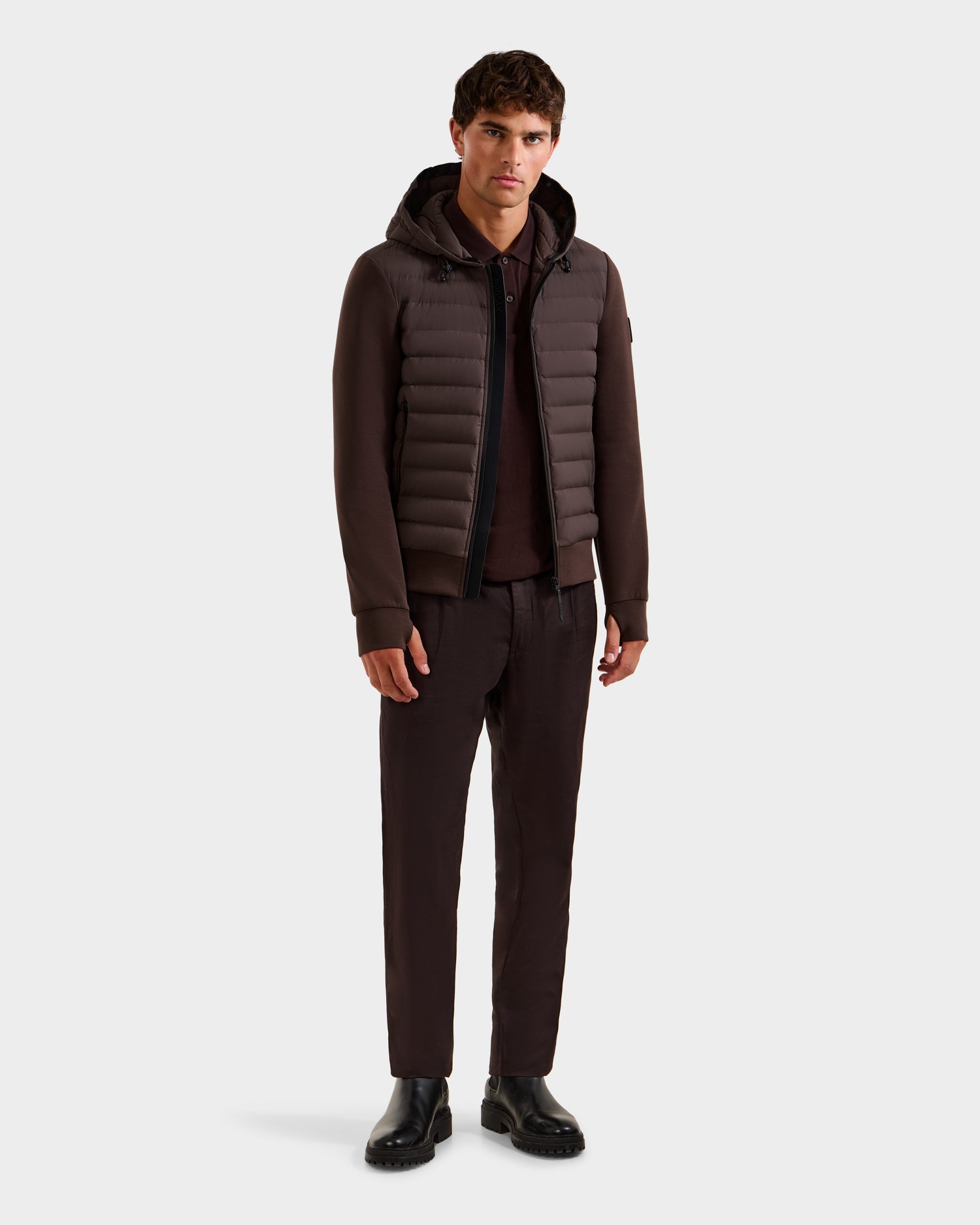 A man stands against a white background wearing the Rudsak DEE MAT - 6124028 Espresso lightweight down jacket, brown pants, a brown shirt, and black shoes.