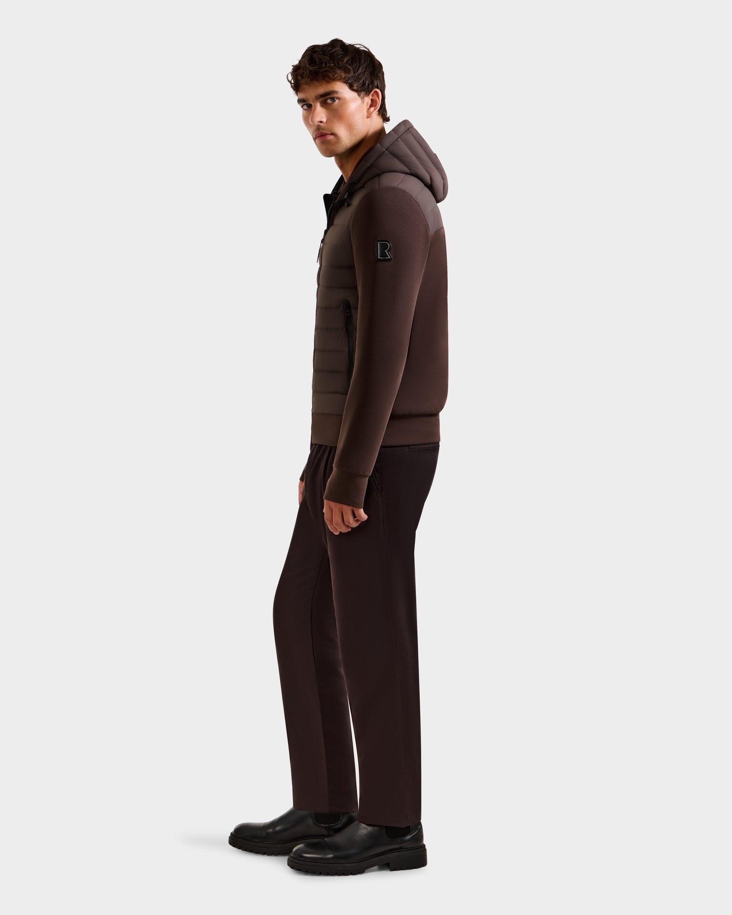 A man stands facing slightly sideways, wearing the Rudsak DEE MAT - 6124028 Espresso hybrid down jacket with a hood, dark brown pants, and black shoes against a plain white background.