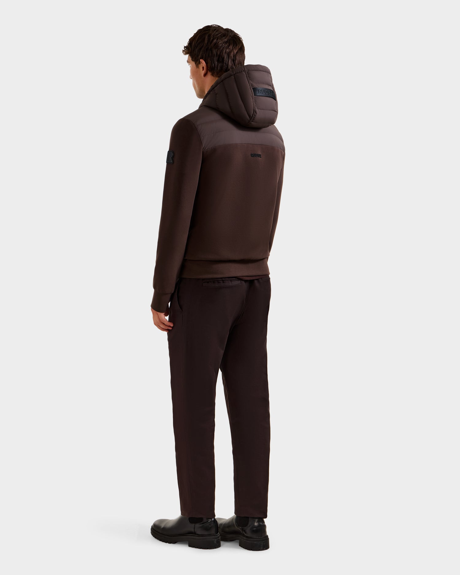 A man faces away, wearing the Rudsak DEE MAT - 6124028 Espresso men's lightweight down jacket with matching pants and black shoes, set against a plain light background.