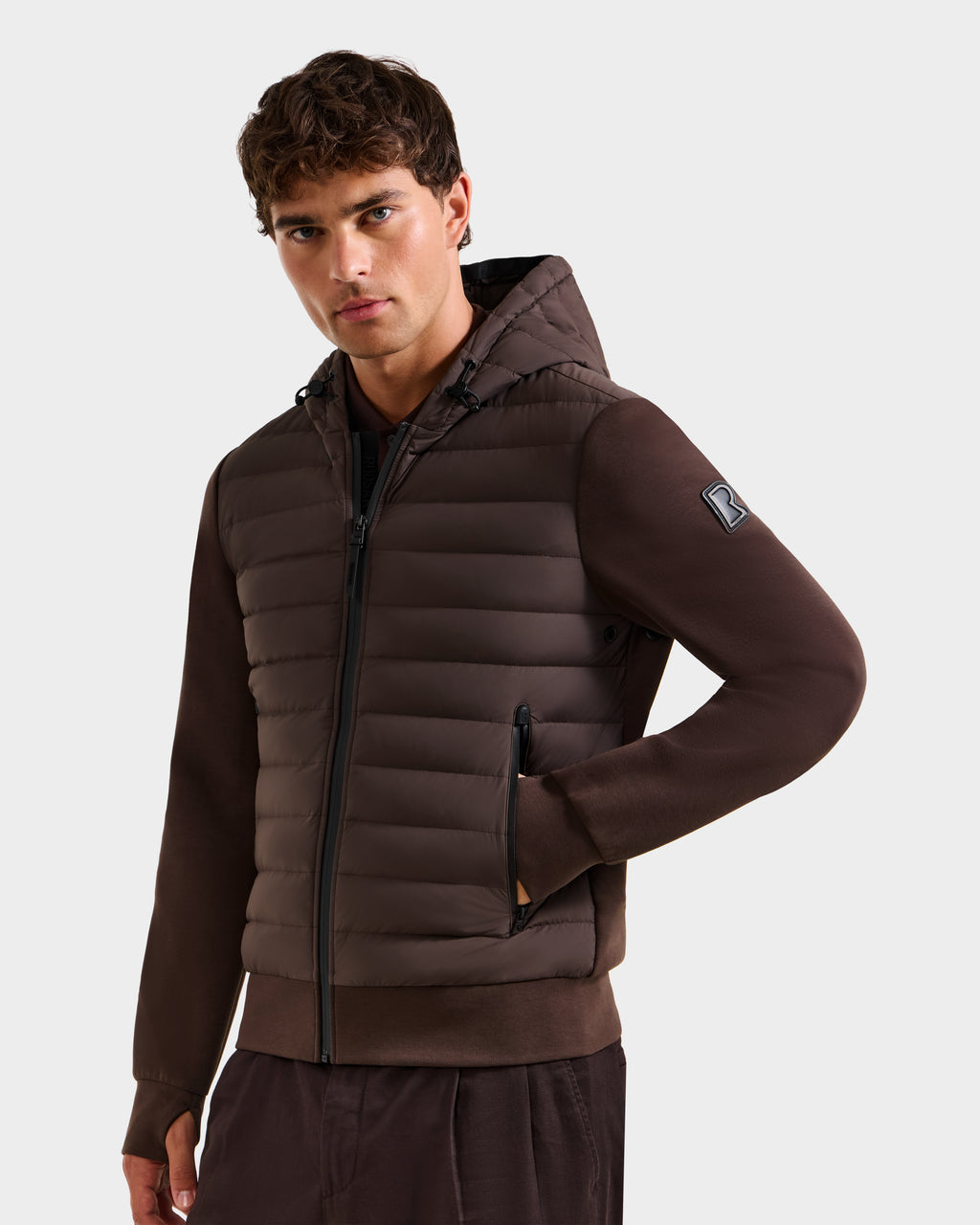 A young man with short brown hair wears the Rudsak DEE MAT - 6124028 Espresso lightweight hybrid down jacket with zippered pockets and matching brown pants, posing with one hand in his pocket against a plain light background.