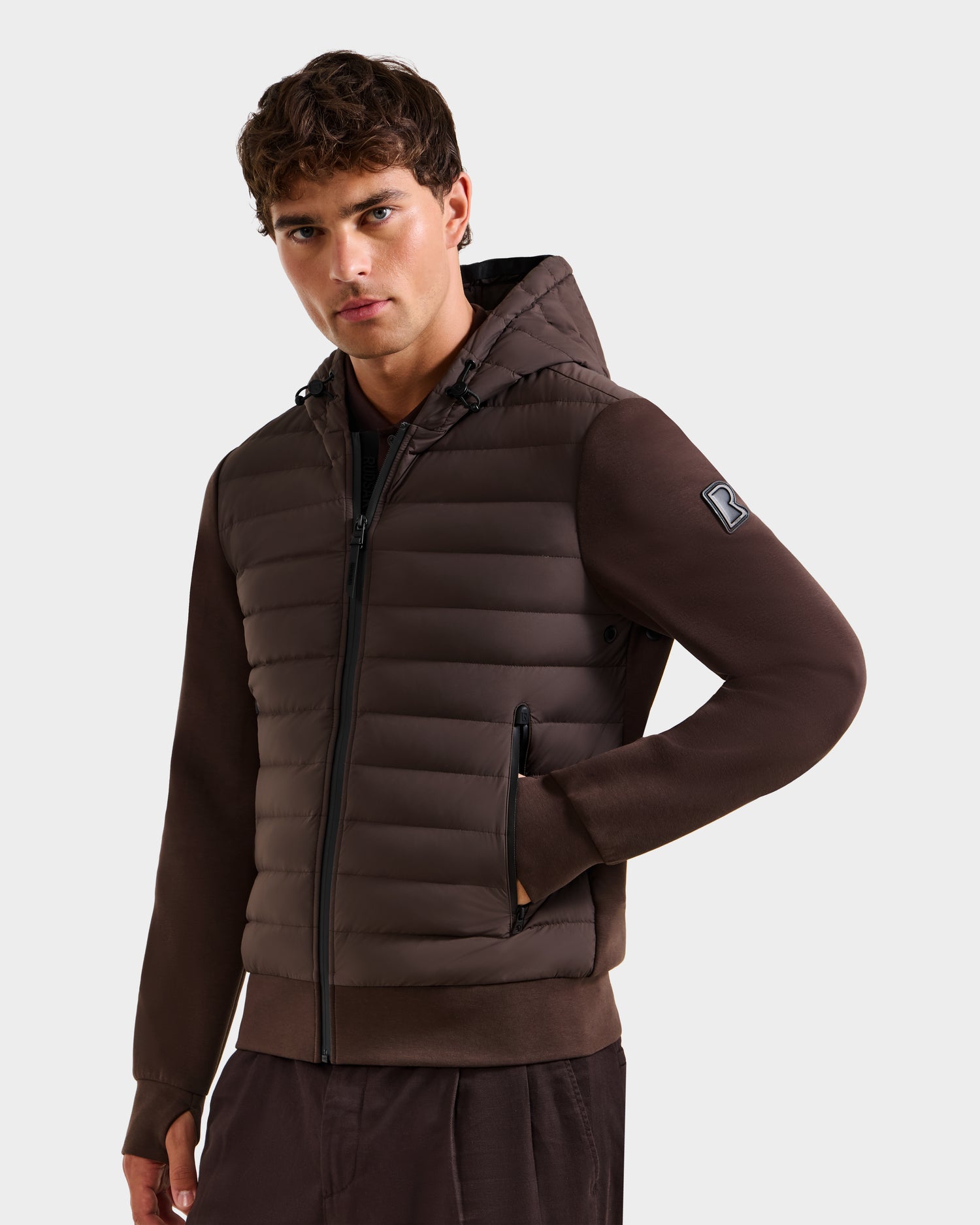 A young man with short brown hair wears the Rudsak DEE MAT - 6124028 Espresso lightweight hybrid down jacket with zippered pockets and matching brown pants, posing with one hand in his pocket against a plain light background.
