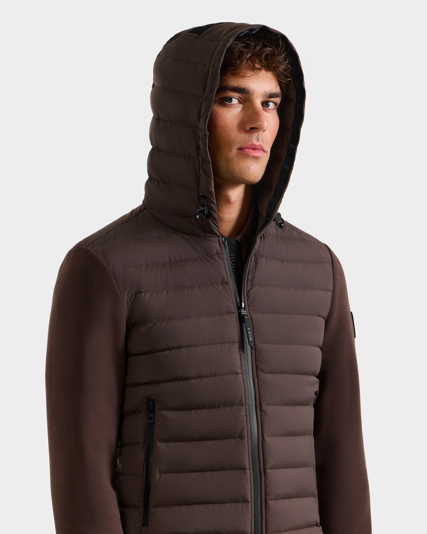 A man wears the Rudsak DEE MAT - 6124028 Espresso urban jacket, featuring a brown padded design, hood, and black zippers, standing against a white background with a neutral expression.