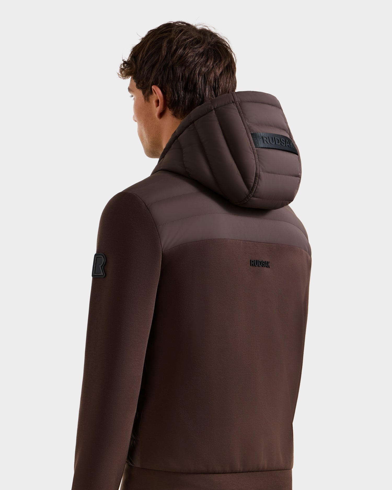 A man is shown from the back and side wearing the Rudsak DEE MAT - 6124028 Espresso lightweight down jacket, highlighting its padded hood, shoulder details, and visible branding on the hood and upper back.