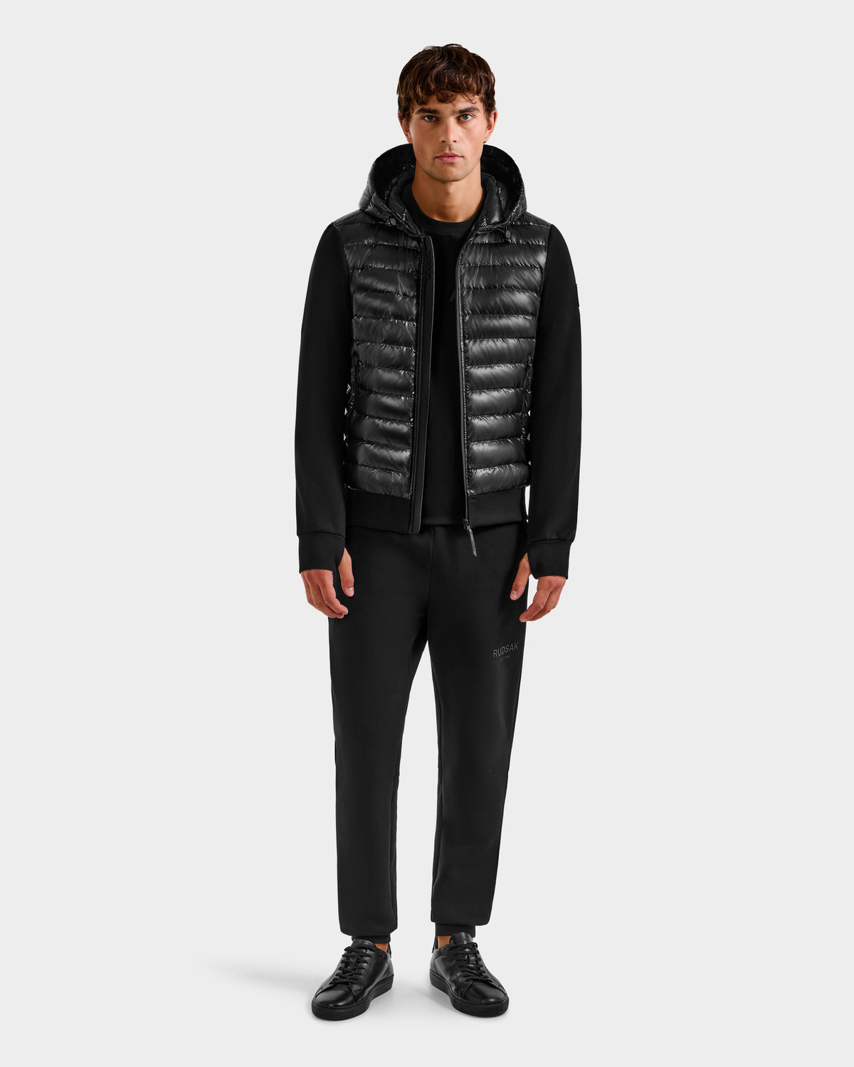 A man faces forward against a plain white background, wearing Rudsak's DEE S - 6125684 Black quilted vest over a black hoodie, paired with black pants and black sneakers.