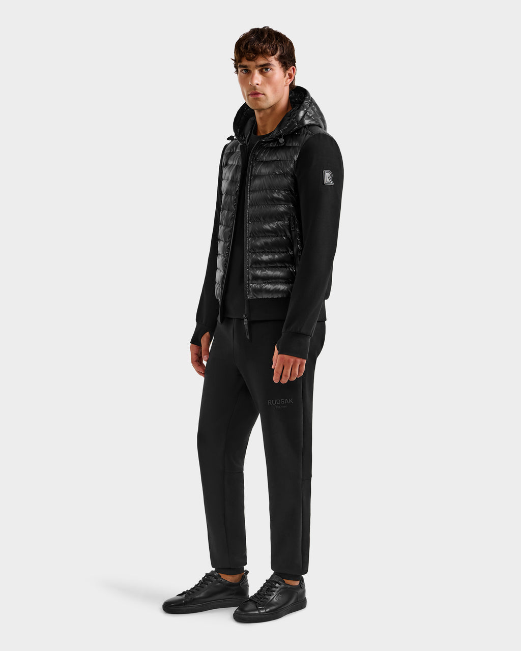 A man stands against a white background wearing the Rudsak DEE S - 6125684 Black hooded puffer vest over a black hoodie, with black joggers and sneakers. He looks slightly to the side, hands by his sides.
