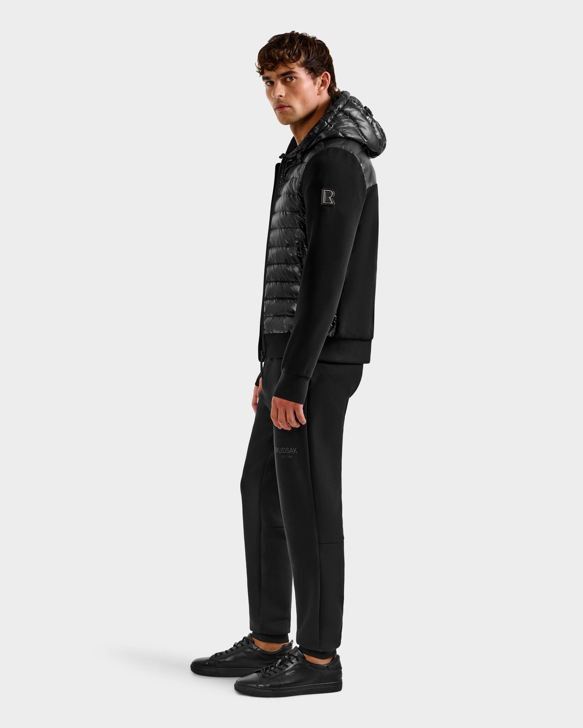 A man stands sideways against a plain white background, wearing the Rudsak DEE S - 6125684 Black men's lightweight down jacket with RDS-certified duck down, paired with black jogger pants and sneakers.
