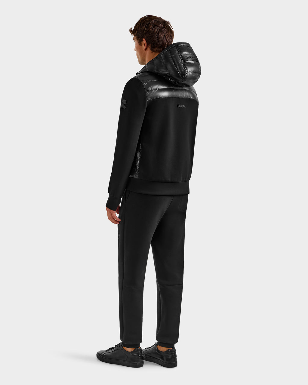 A man faces away wearing Rudsak’s DEE S - 6125684 Black men’s lightweight down jacket with RDS-certified duck down, paired with black pants and sneakers against a plain, light background.