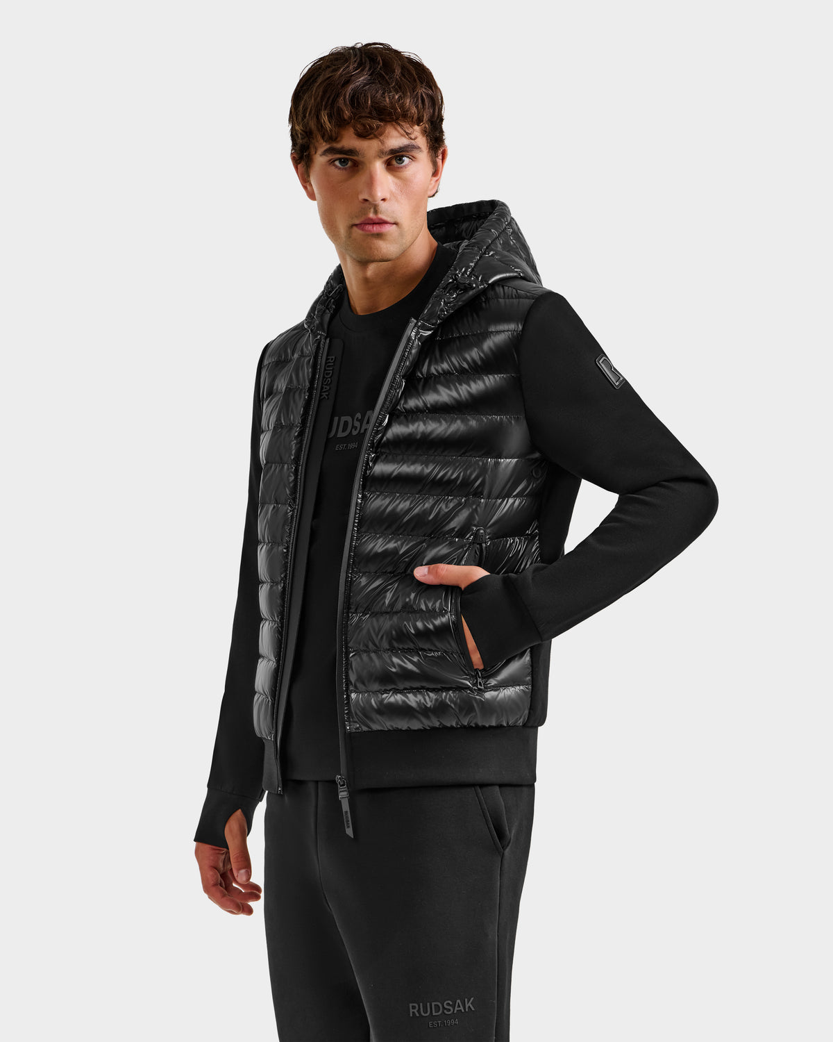 A man stands slightly sideways, wearing the Rudsak DEE S - 6125684 Black—a men’s lightweight, RDS-certified duck down hooded puffer vest—layered over a long-sleeve shirt and pants, embodying effortless urban style with a neutral expression.